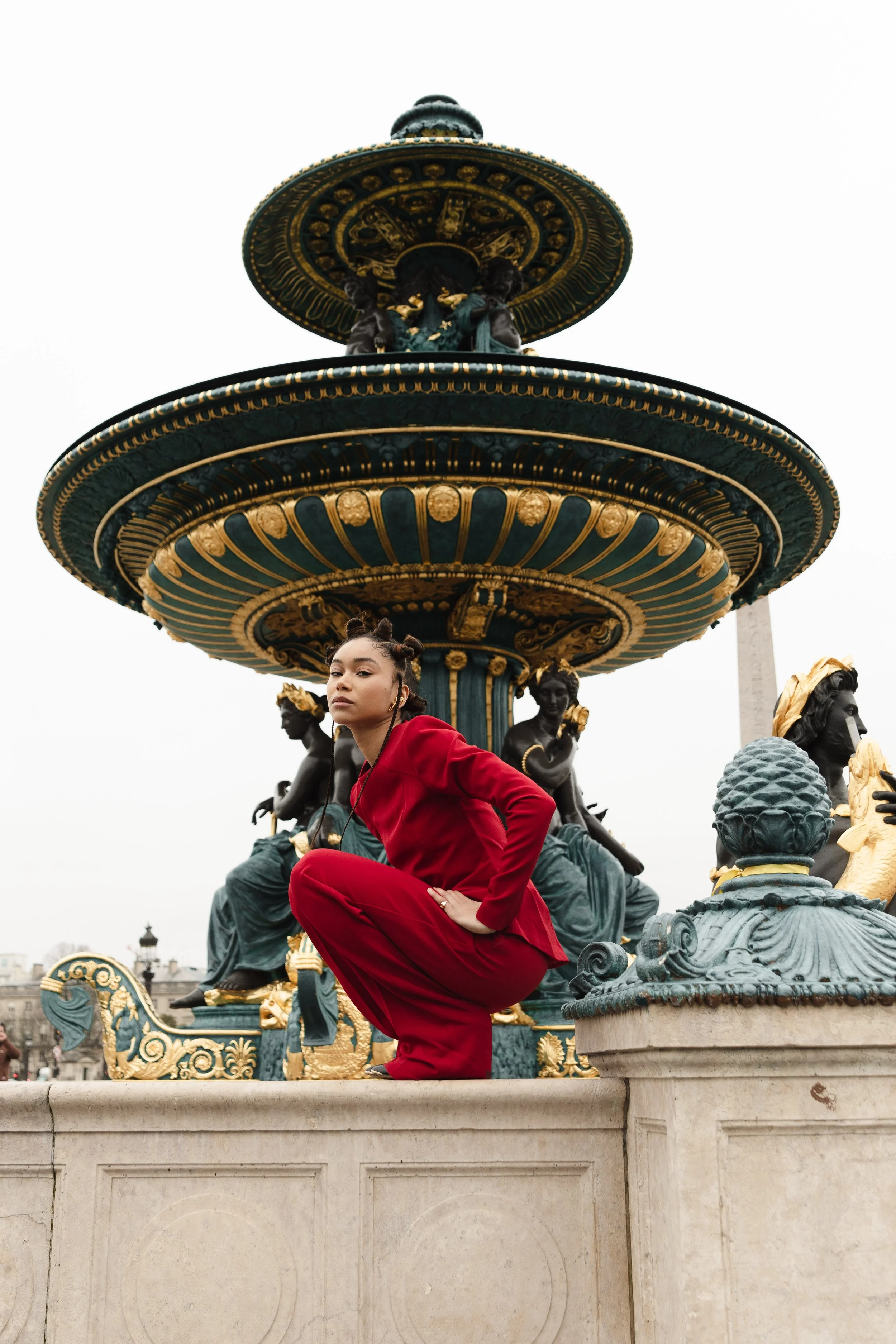 A woman in a red suit poses in front of an ornate fountain with golden decorative elements and black statues, in an outdoor city setting.