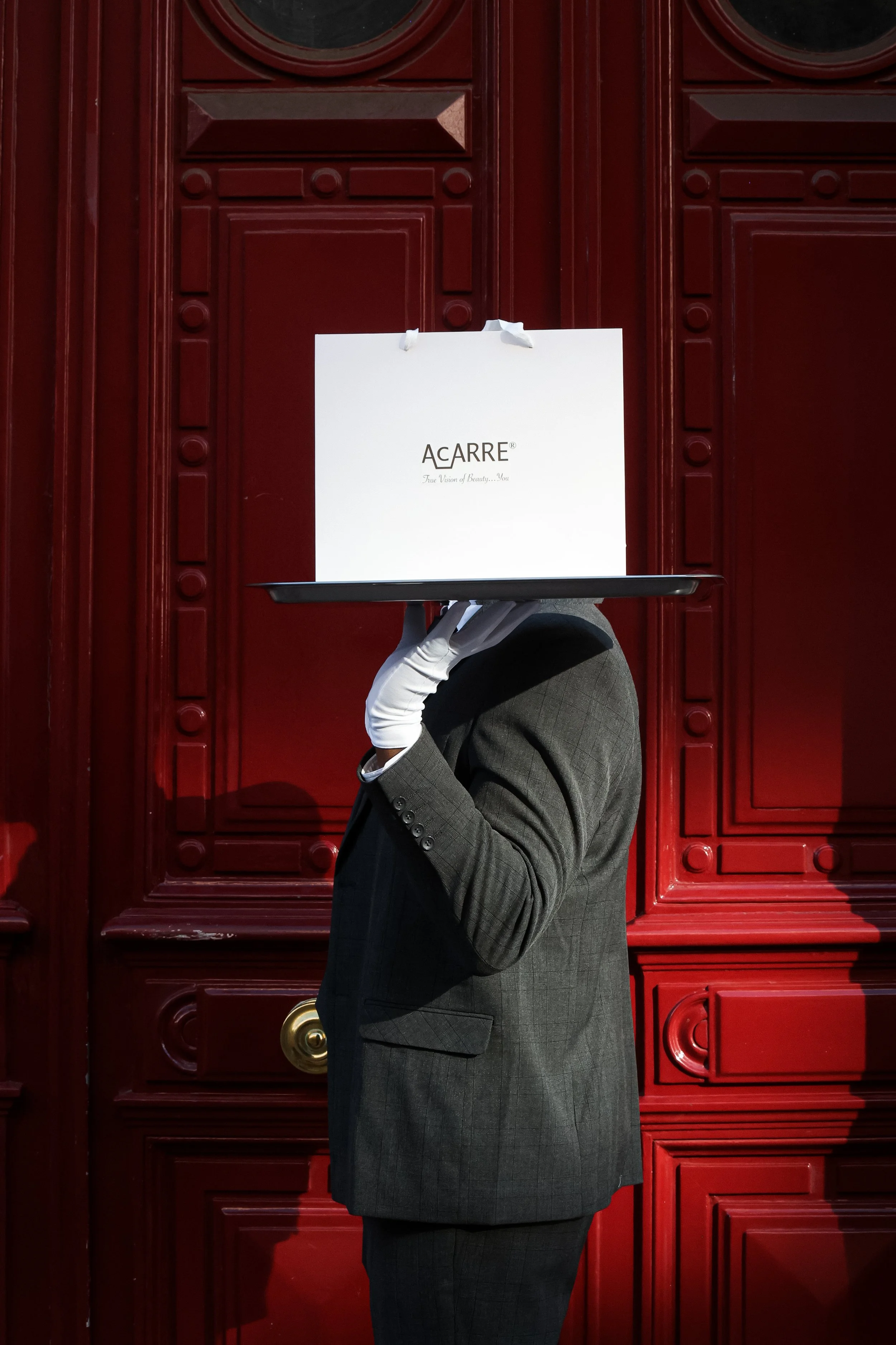 A person in a gray suit, wearing white gloves, holding a large white shopping bag with the text 'AcARRE' in front of a red decorative door.
