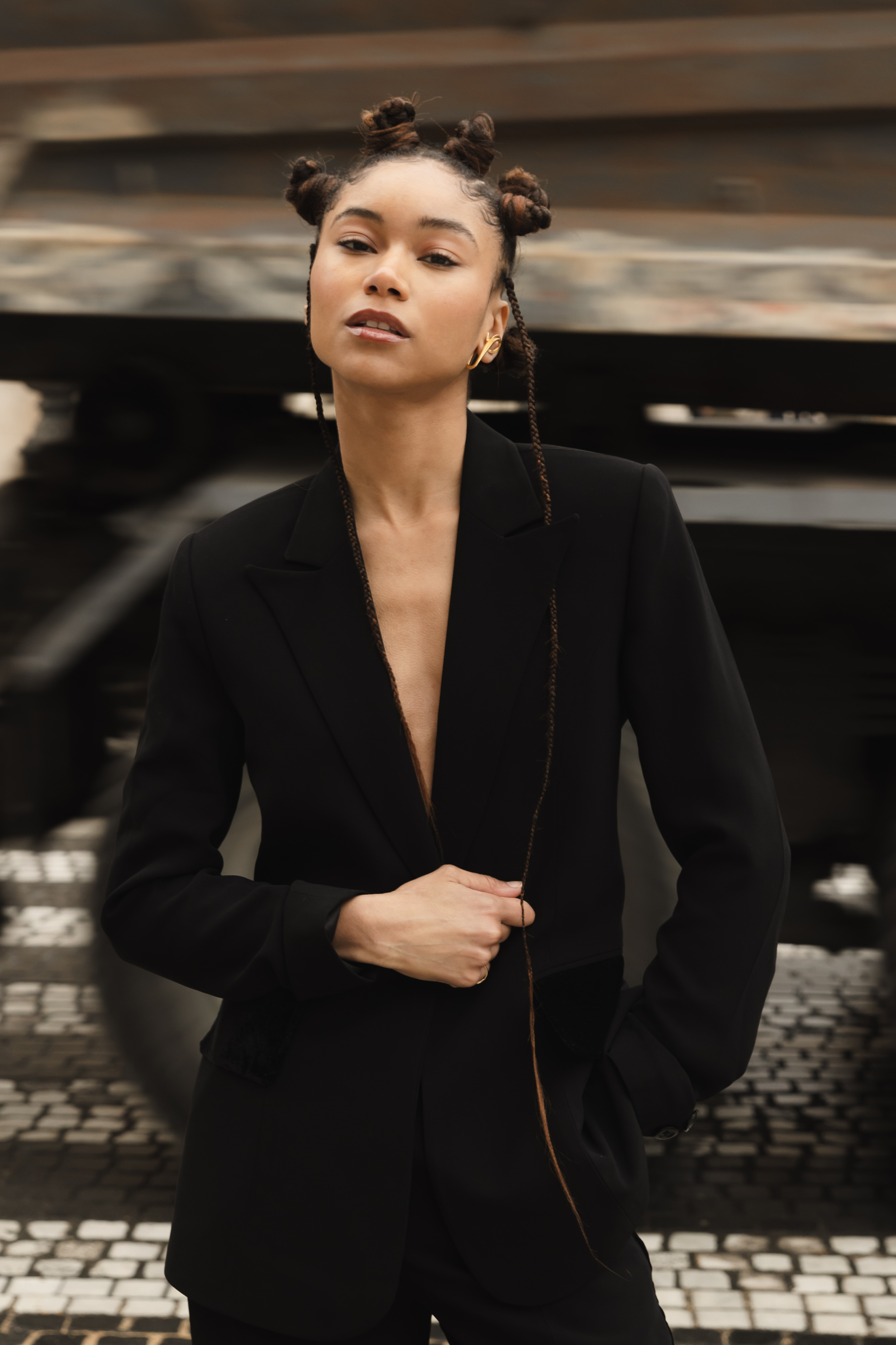 A woman with styled hair and gold earrings wearing a black blazer, standing outdoors with a blurred industrial background.