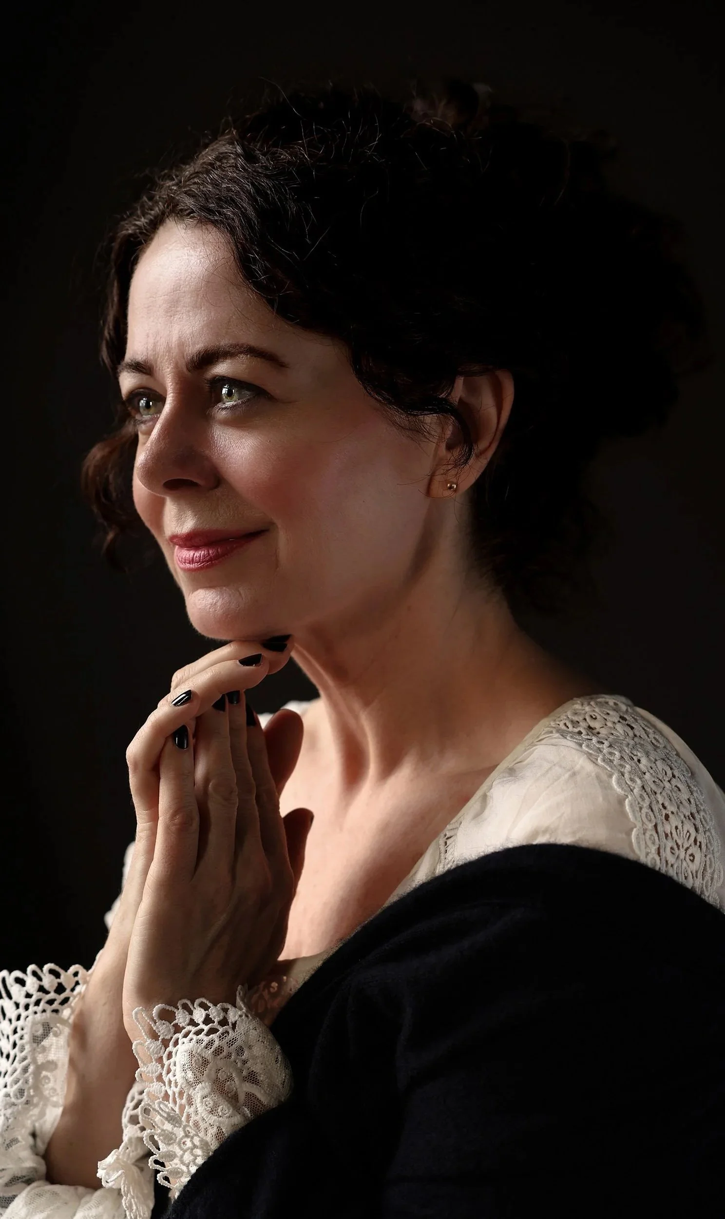 A woman with short, dark curly hair and light skin, wearing a cream-colored lace top with a black outer garment. She is touching her chin lightly with a gentle smile, against a dark background.