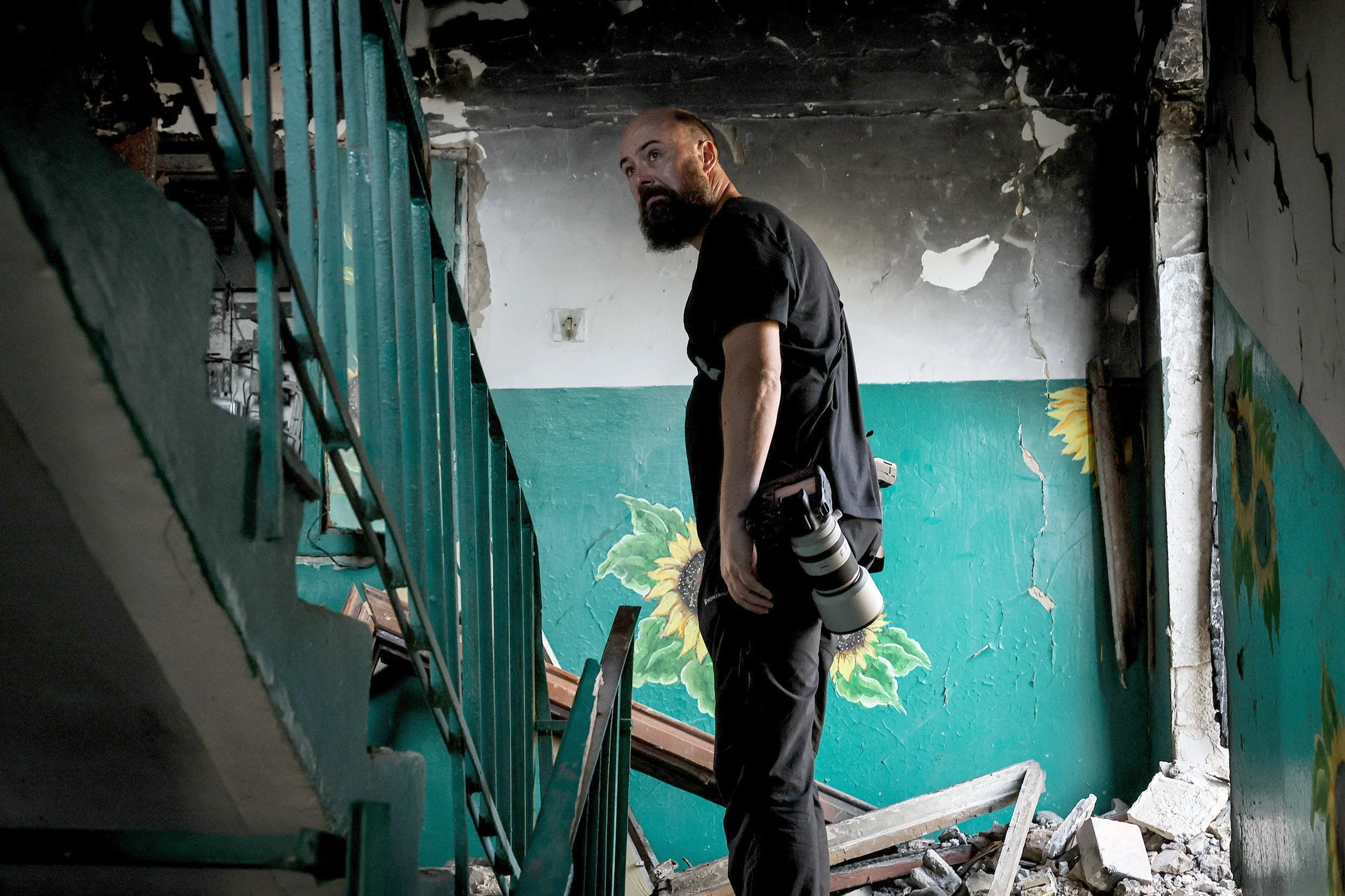 A man with a camera hanging from his shoulder stands in front of a damaged wall with cracks and rubble, inside a building with painted wall murals of sunflowers.