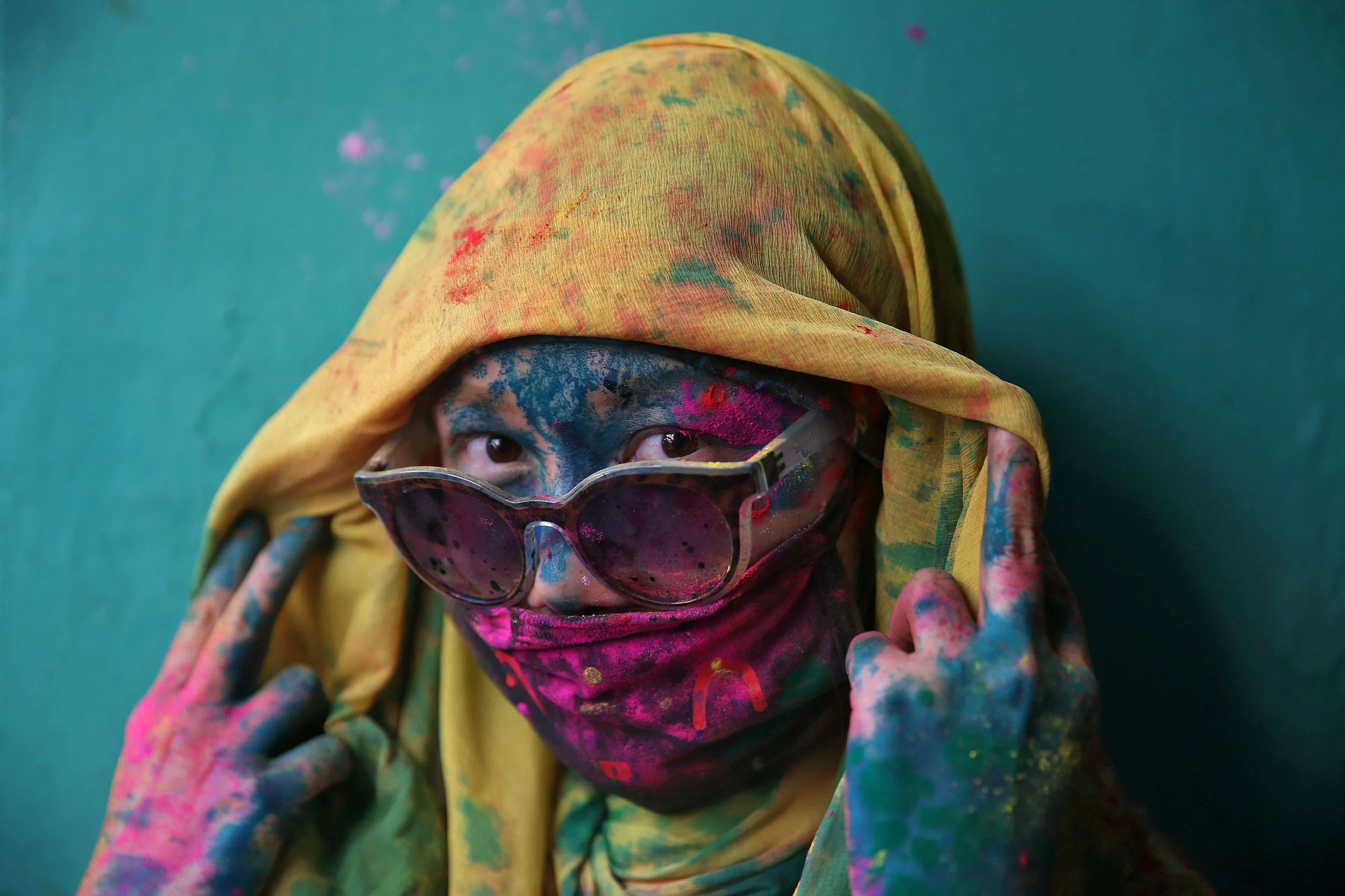 Person with blue, pink, and yellow paint on face and hands, wearing sunglasses and a yellow headscarf, against a teal background.