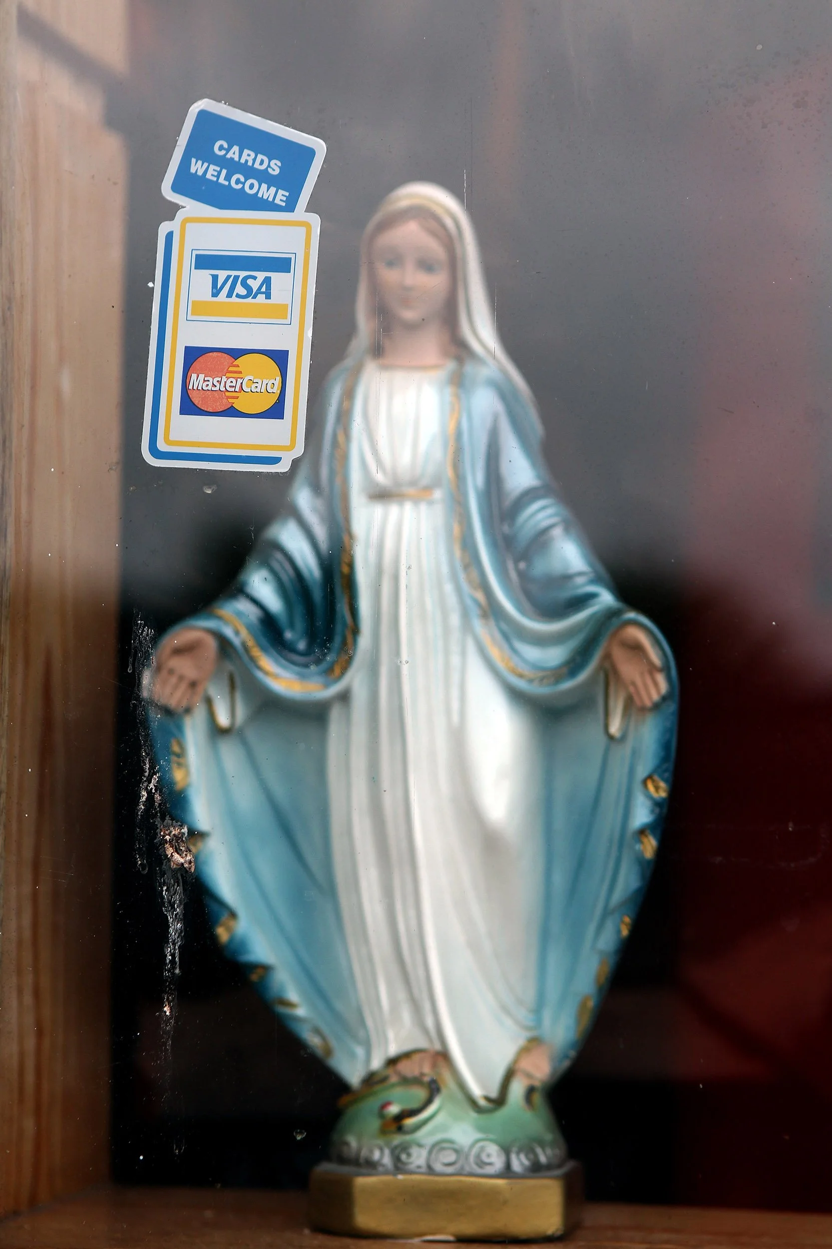 A religious statue of the Virgin Mary, depicted with a white dress and a blue cloak, standing on a decorative base. Stickers on the glass say 'Cards Welcome,' 'VISA,' and 'MasterCard.'