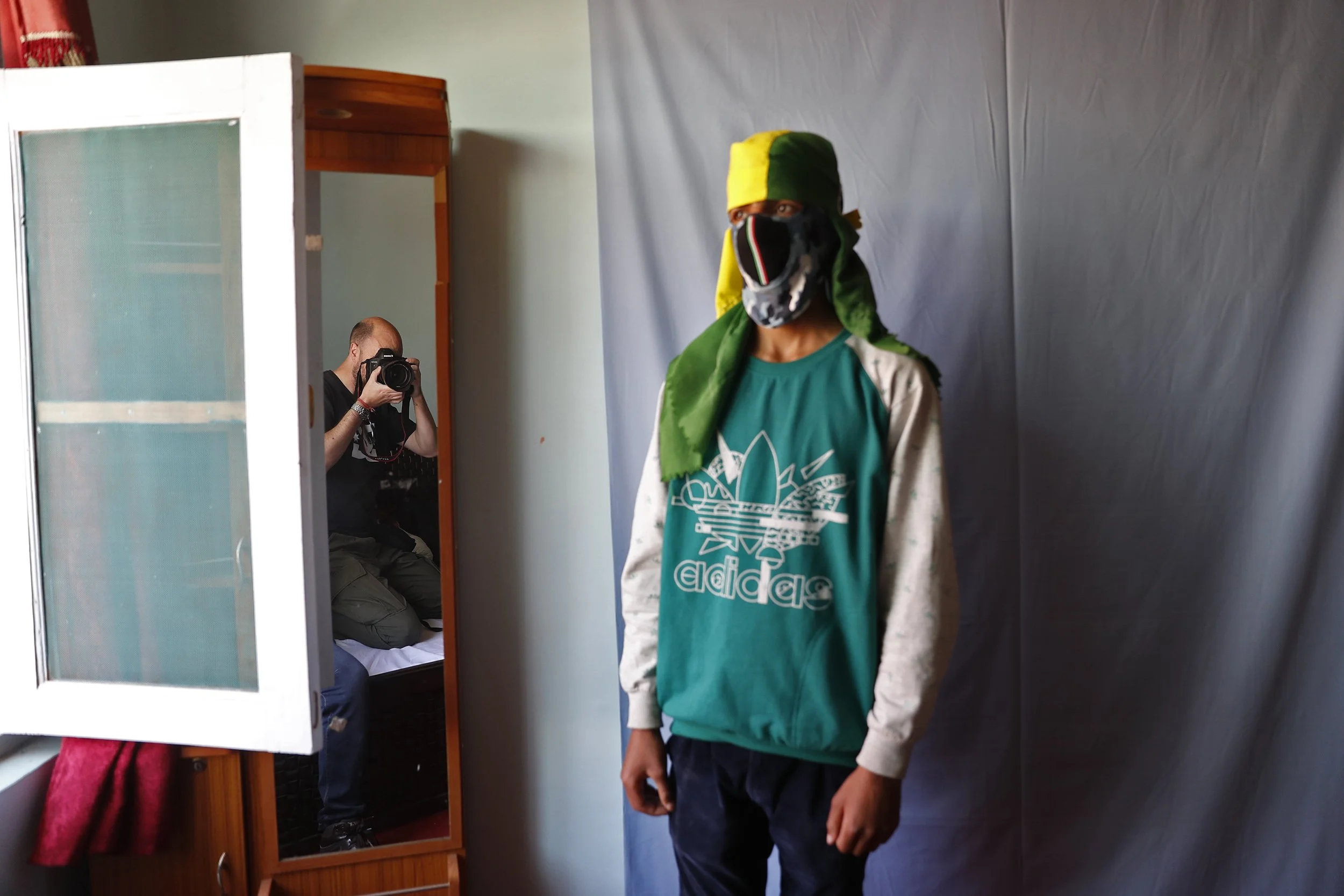 A person wearing a green and yellow head covering, a black face mask, a teal Adidas shirt, and white hoodie standing next to a gray backdrop, with a photographer taking their picture in a mirror.