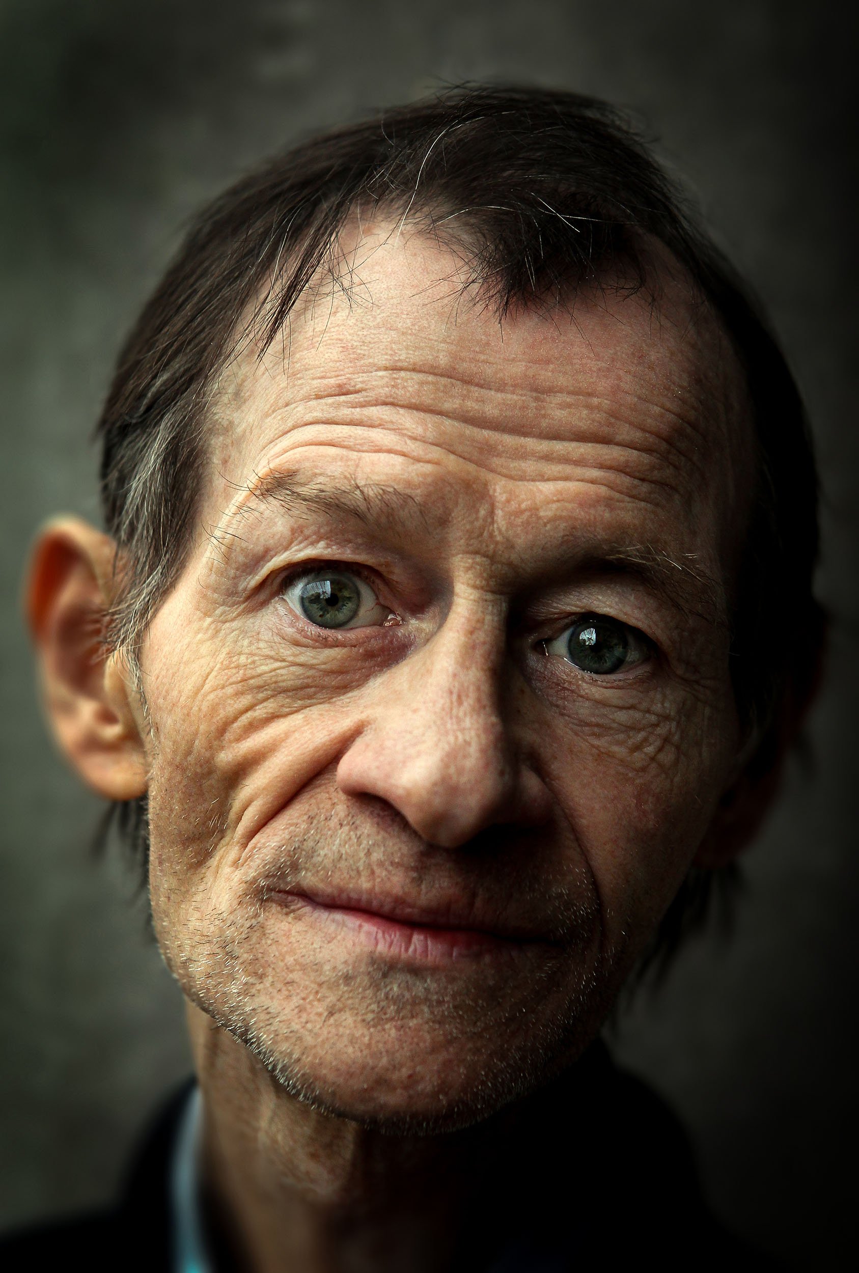 A close-up portrait of an elderly man with blue eyes, wrinkles, and stubble, looking directly at the camera against a dark background.