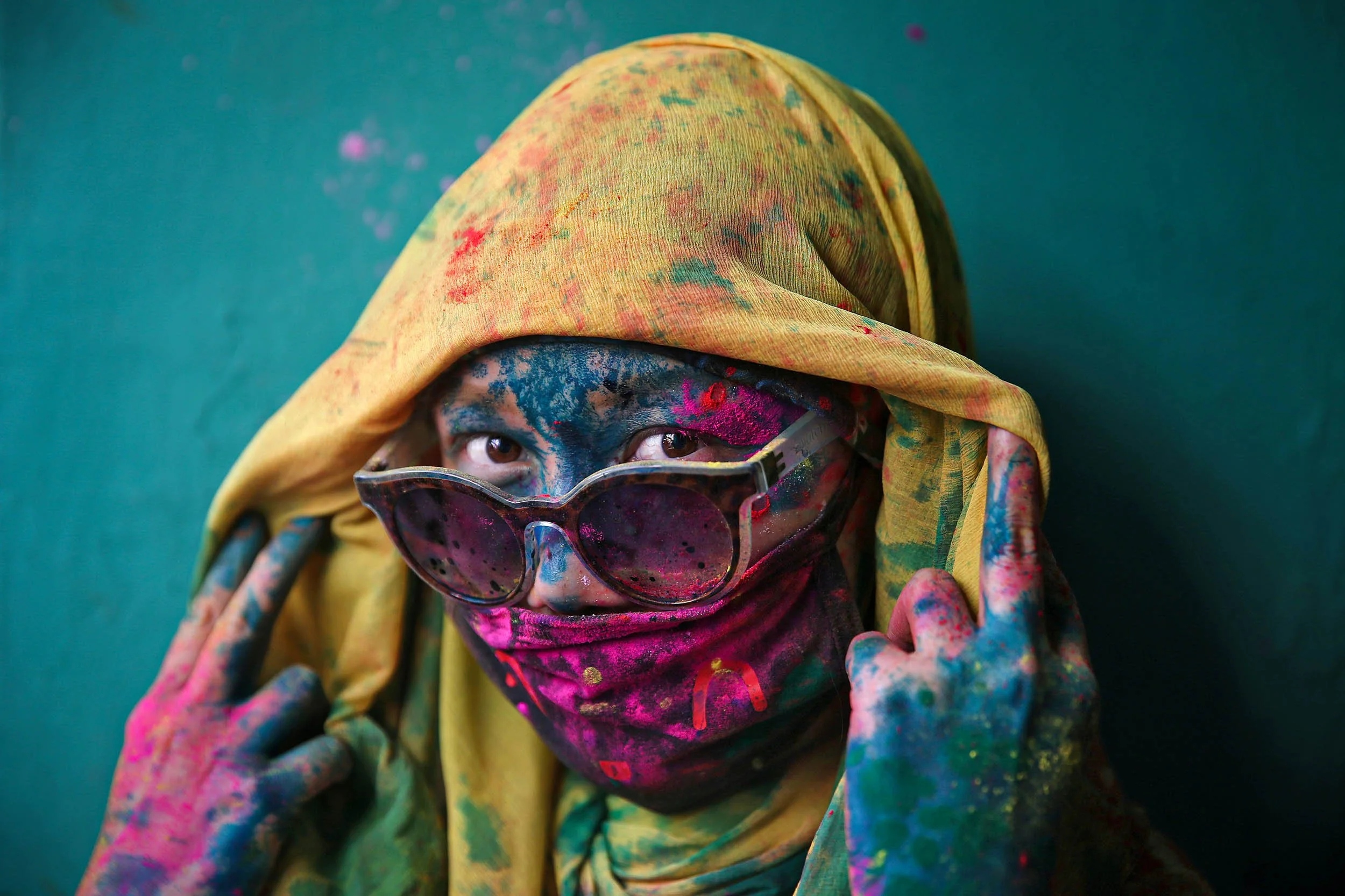 Person with paint-splattered face, wearing pink sunglasses, yellow headscarf, and multicolored paint-stained hands, against a teal background.