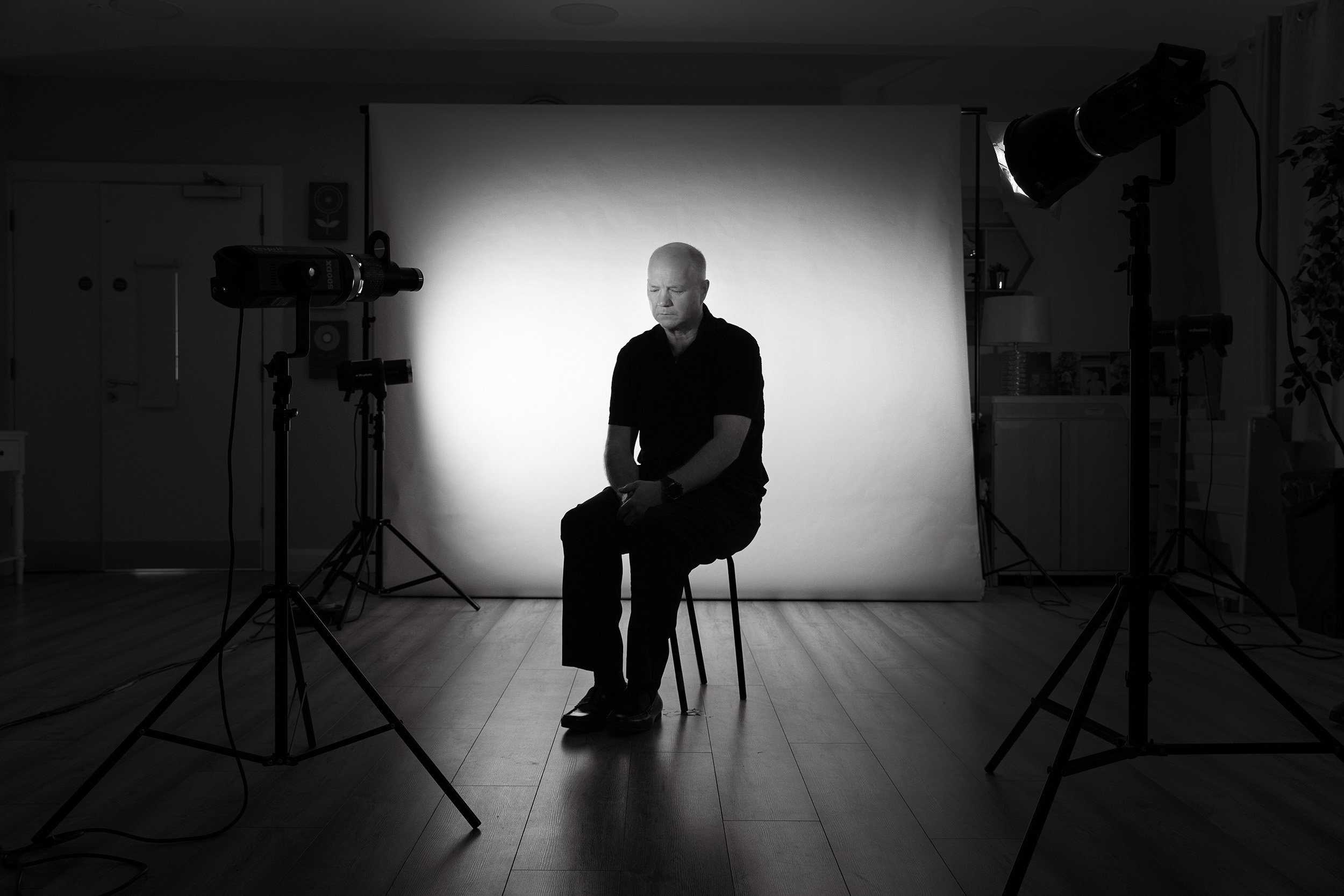A person with a shaved head sitting on a stool in a photography studio, surrounded by studio lights and a backdrop.