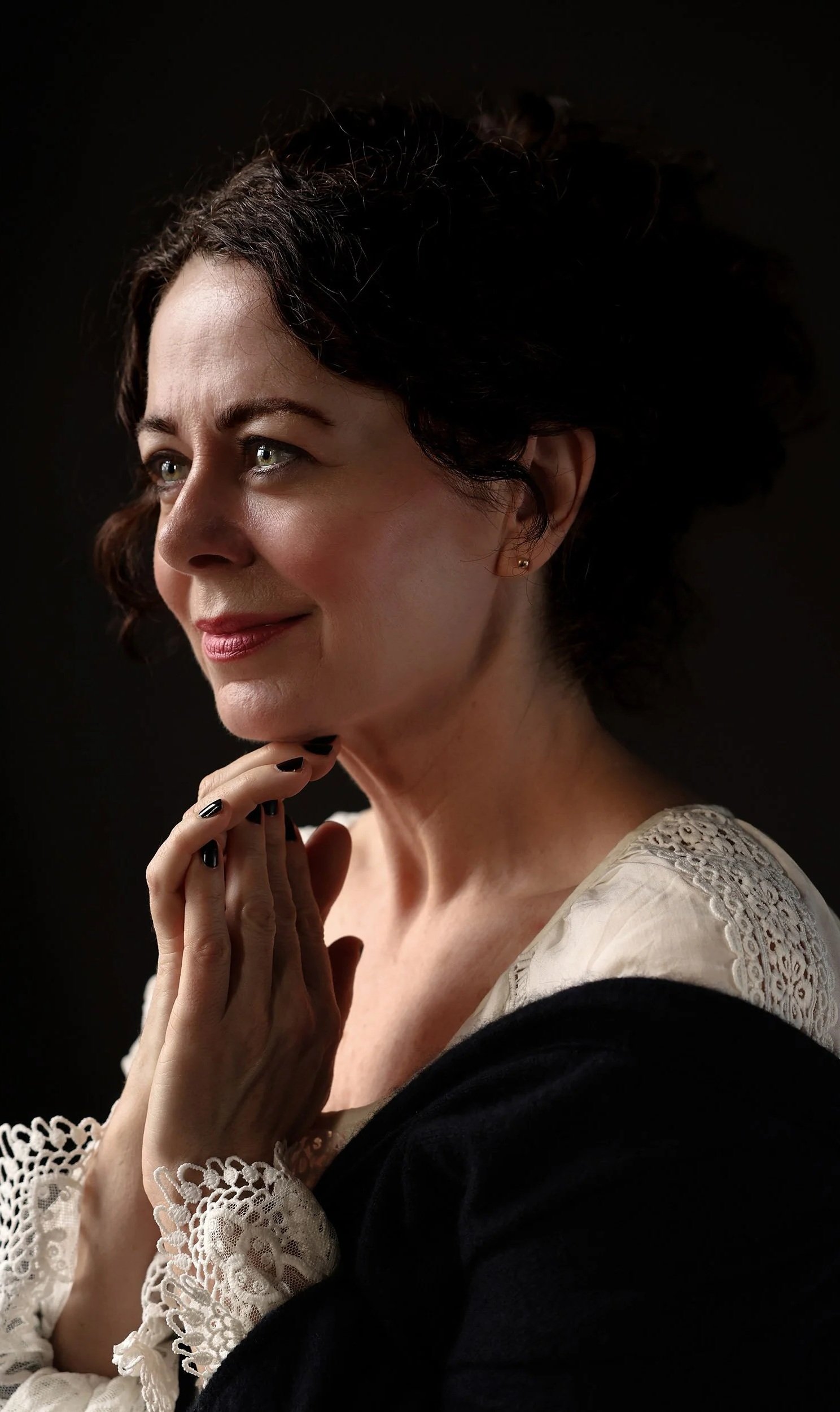 A woman with dark curly hair and light skin, wearing a cream-colored top with lace details, poses thoughtfully with her hands gently resting near her chin, against a dark background.