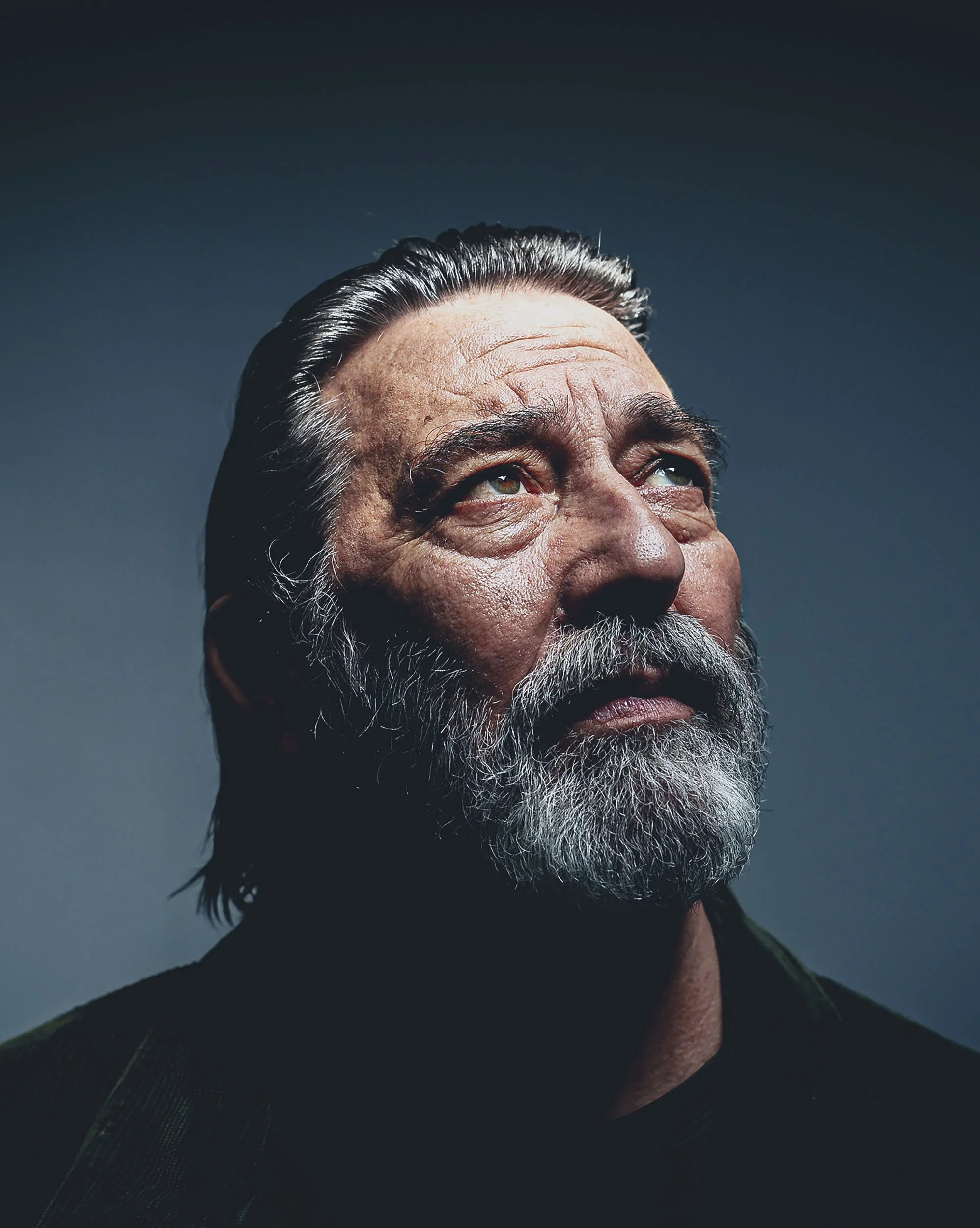 Close-up portrait of an elderly man with a gray beard and mustache, looking upwards against a dark background under soft lighting.