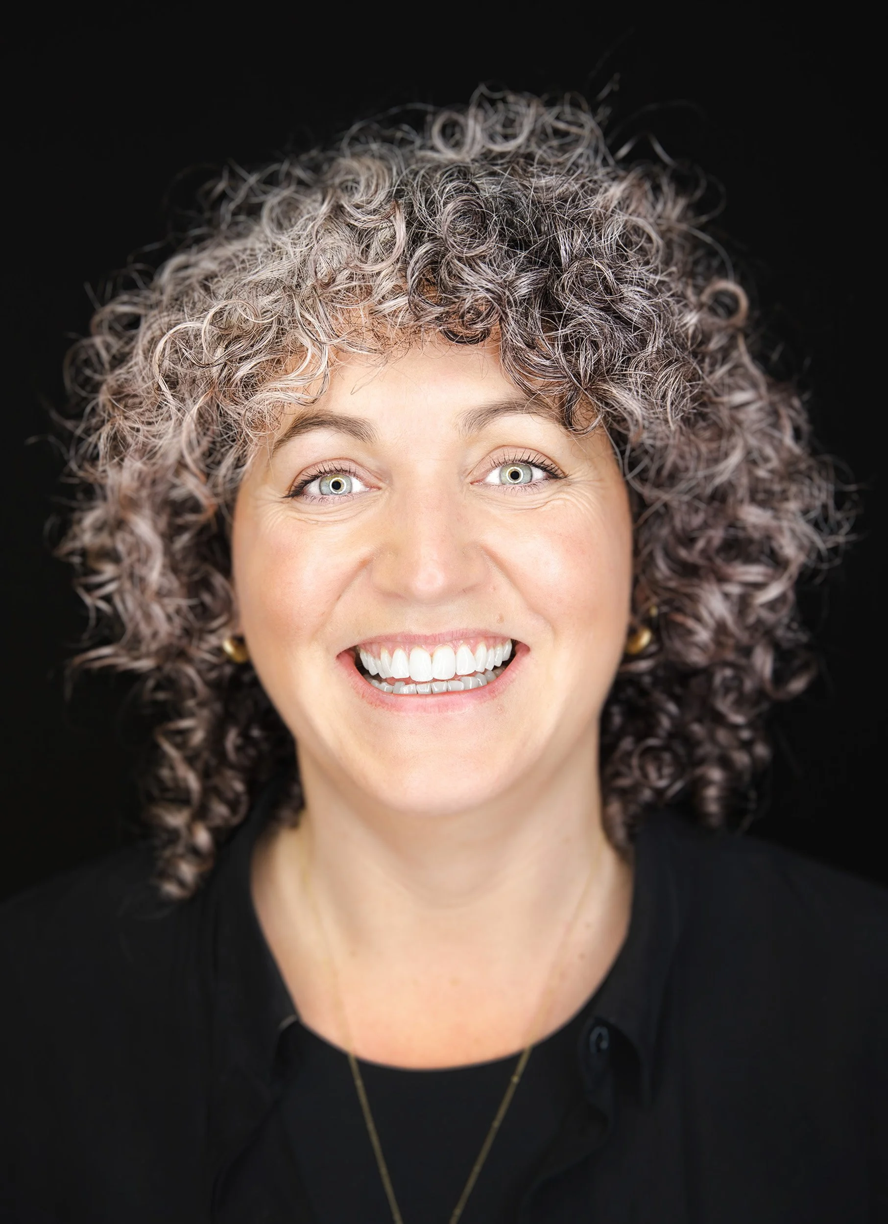 A woman with curly gray hair and bright eyes smiling at the camera, wearing a black top and a gold necklace, against a black background.