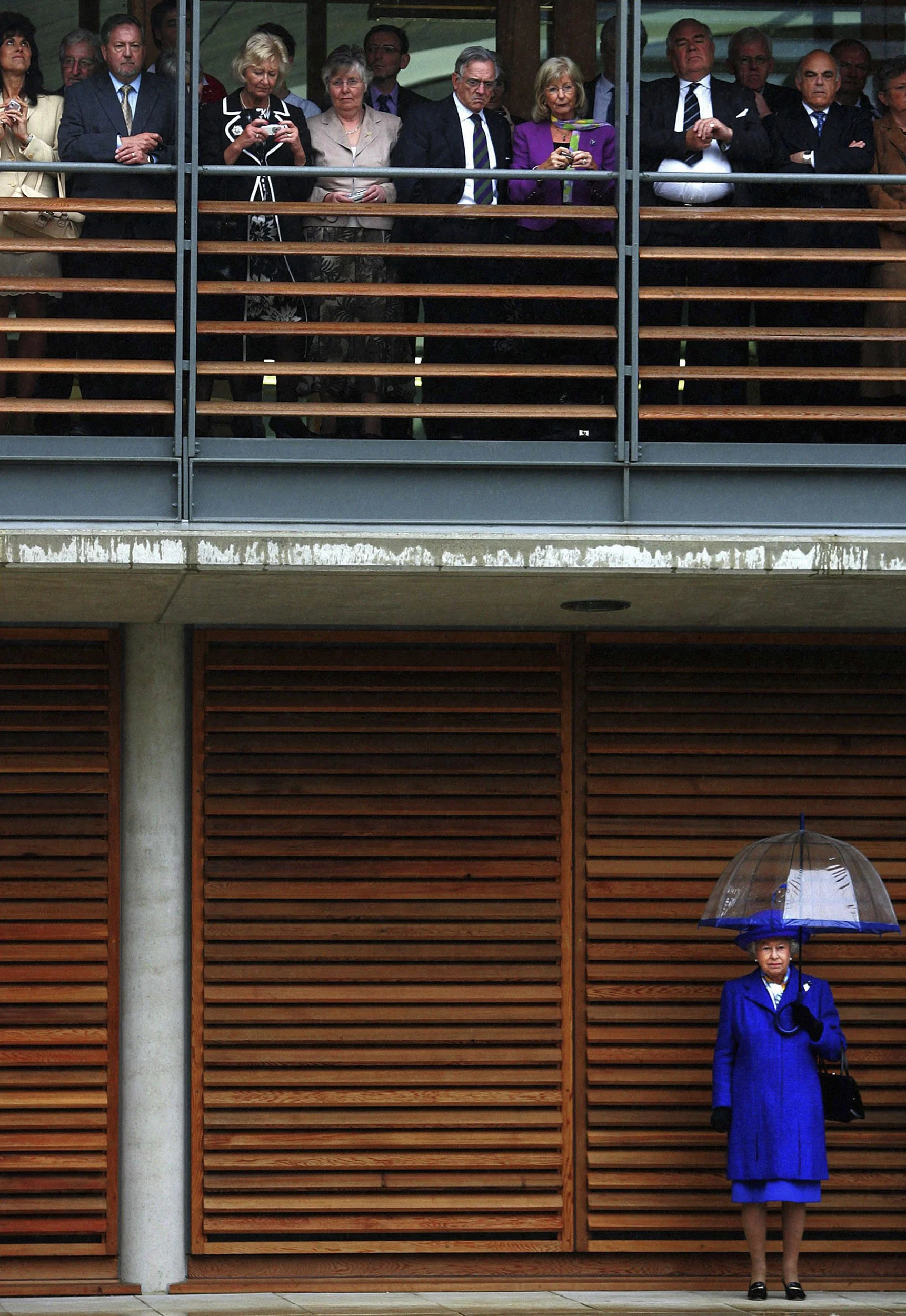 A group of people on an upper balcony, some taking photos, viewing an event below. A woman on the ground, dressed in blue with black shoes, holds a transparent umbrella, standing against a wooden wall.