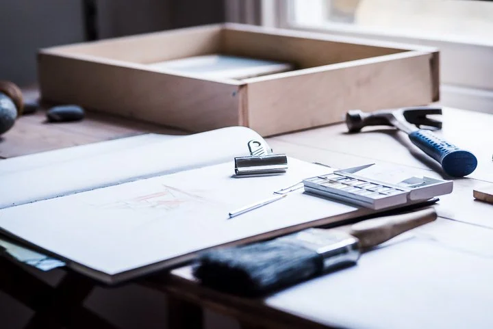 Sketch book, paints and a hammer lay on designers desk, showing designs being produced