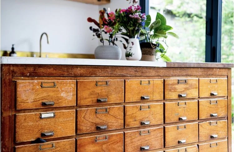 A vintage haberdashery unit re purposed as a kitchen island with topped with a bespoke concrete worktop, decorated with colourful flowers and green plants in vases, and positioned near a window letting in natural light.