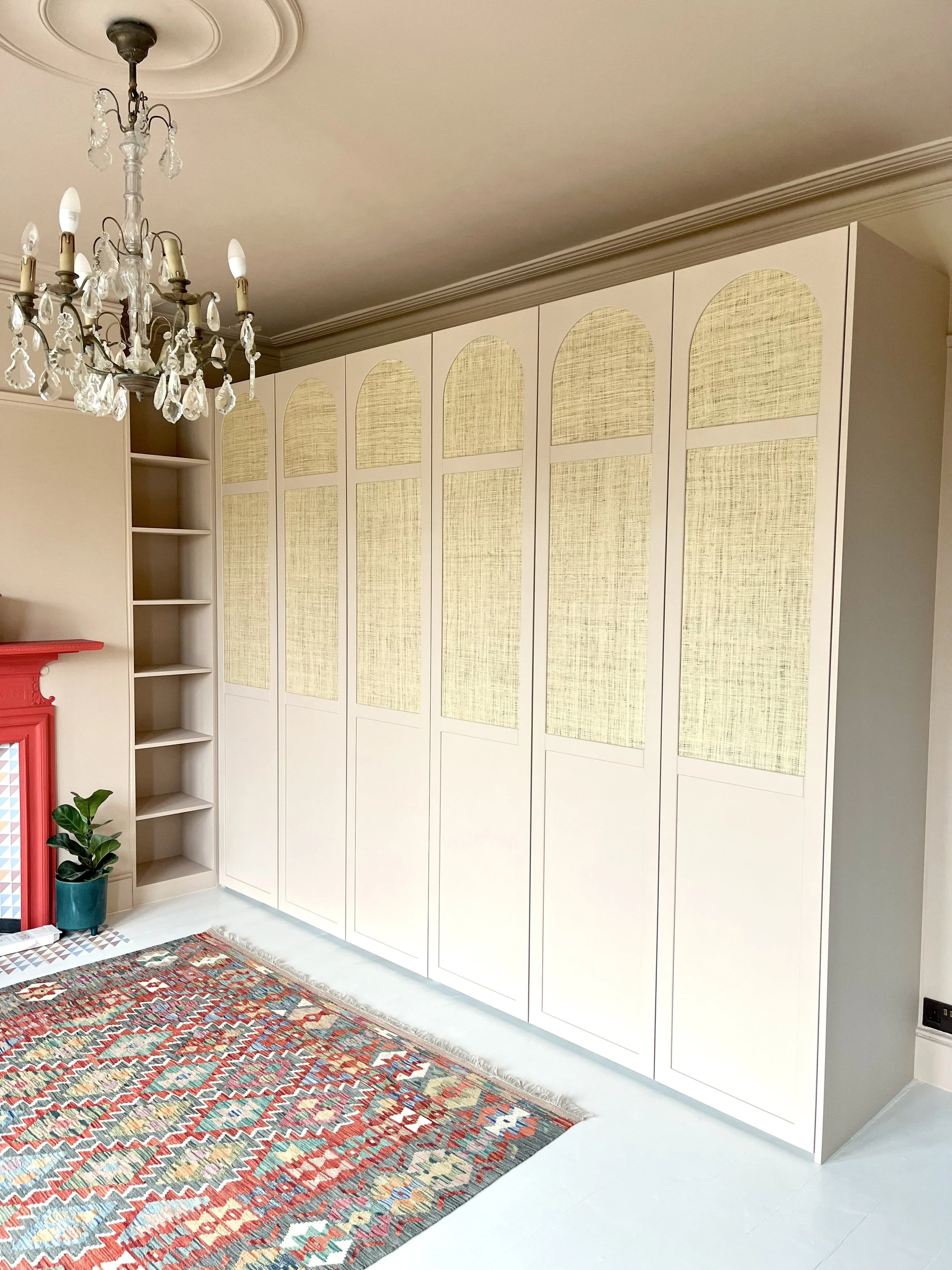 A room with a large white built-in wardrobe featuring arched yellow textured panels on the doors, a white ceiling with decorative molding, a chandelier with hanging crystal accents, a colorful patterned rug, a small potted plant, and a size with red and blue accents.