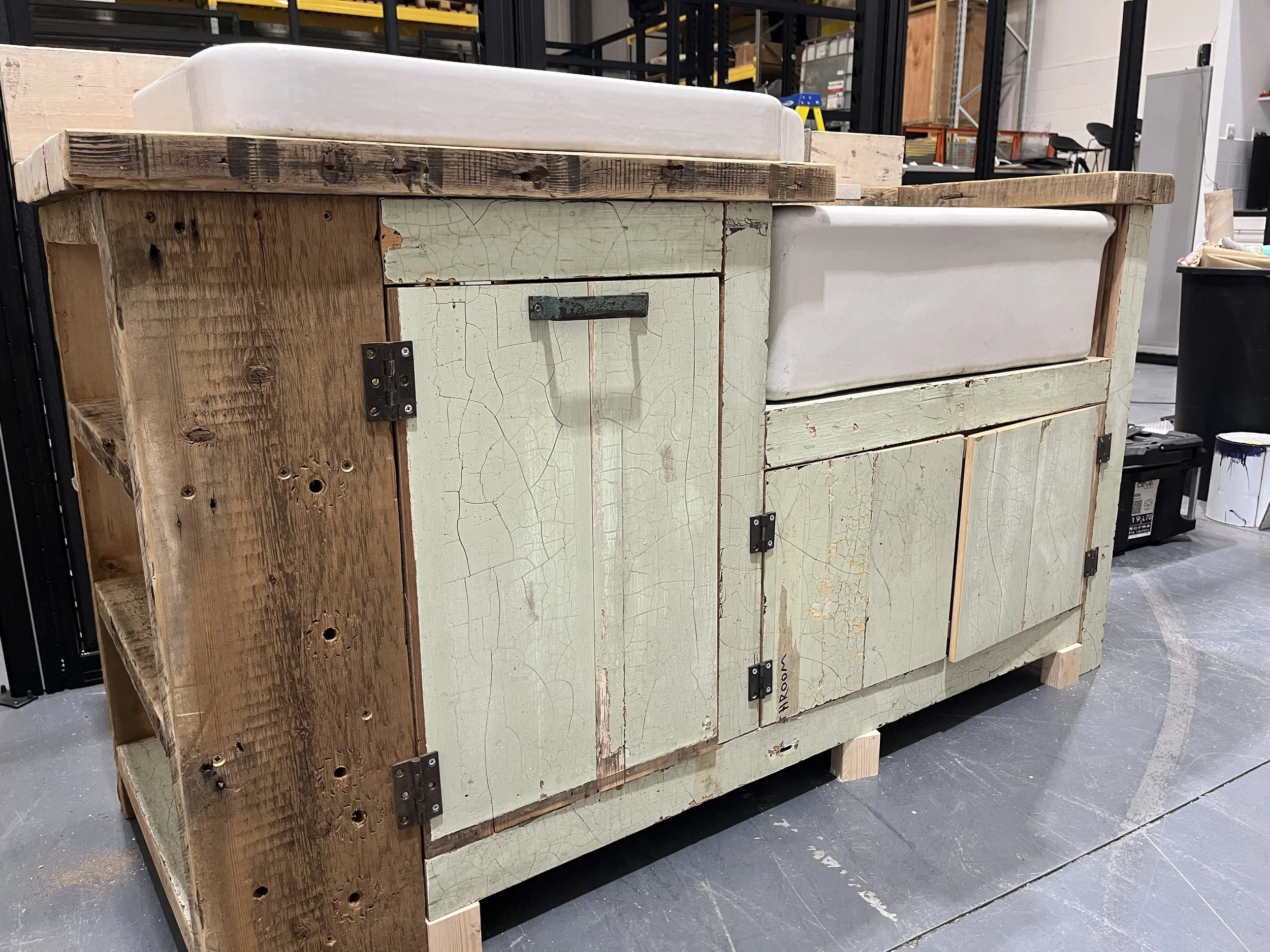 A rustic wooden cabinet with a distressed light green painted finish, showing cracks and wear, housing a vintage farmhouse sink with a white basin, situated in a workshop or garage setting.