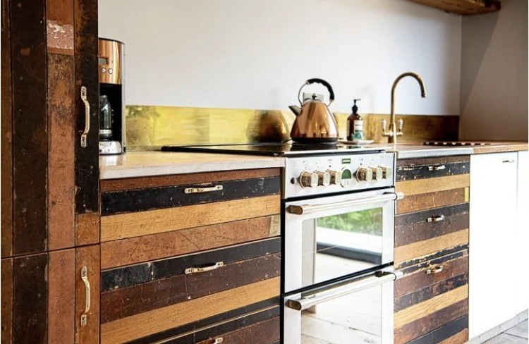 Kitchen with rustic wooden cabinets, a white stove with control knobs, a copper kettle, a soap dispenser, a sink with a faucet, and a window reflected on the oven door