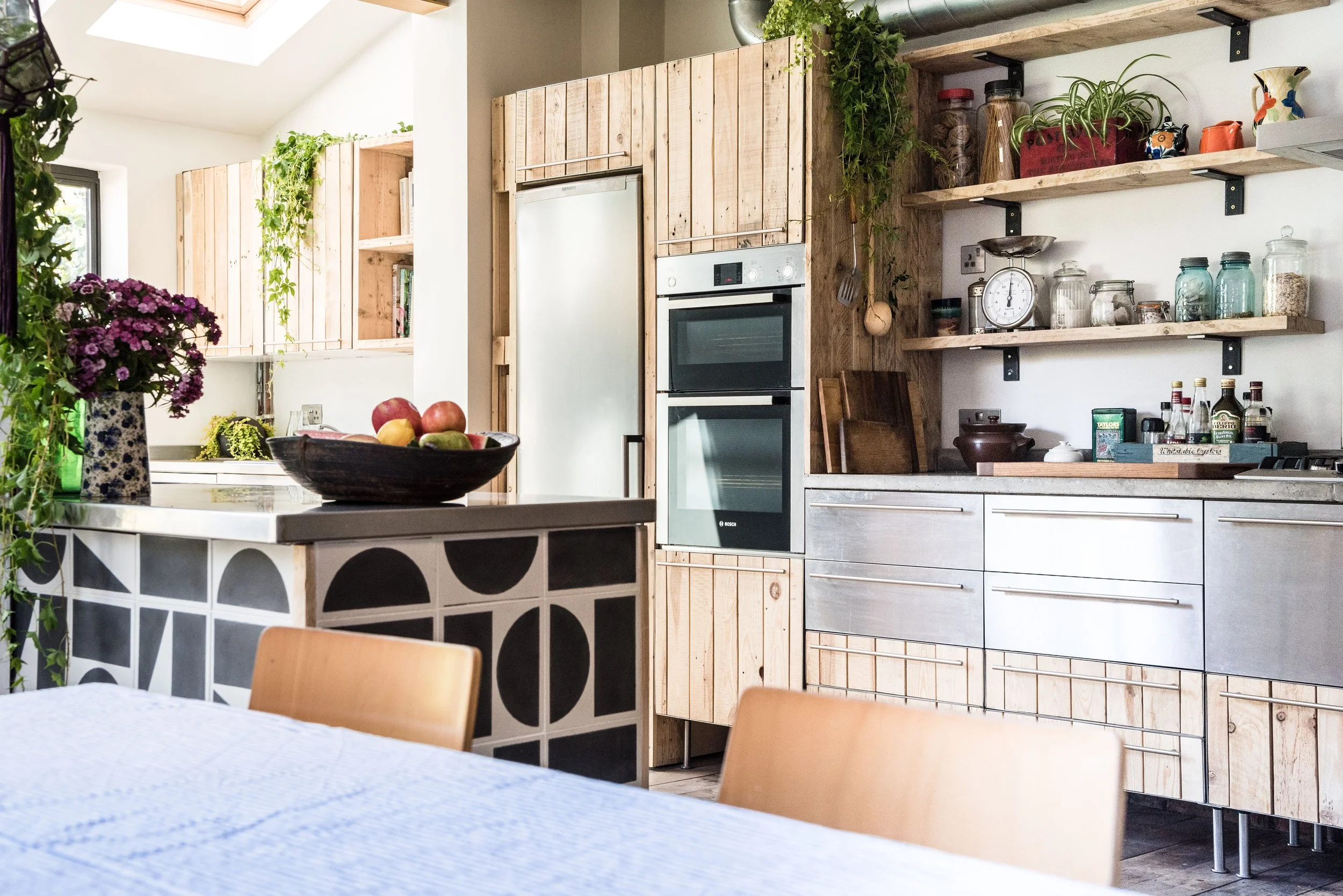 Modern kitchen with wooden cabinets, open shelves with jars and plants, and a dining table with chairs. A bowl of apples is on the counter, and sunlight streams through a skylight.