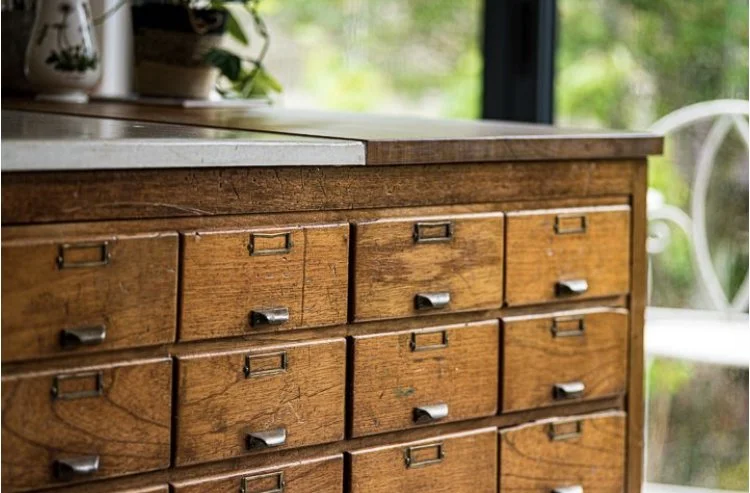 Close-up of a vintage wooden cabinet with multiple small drawers and metal handles, near a window with greenery outside.