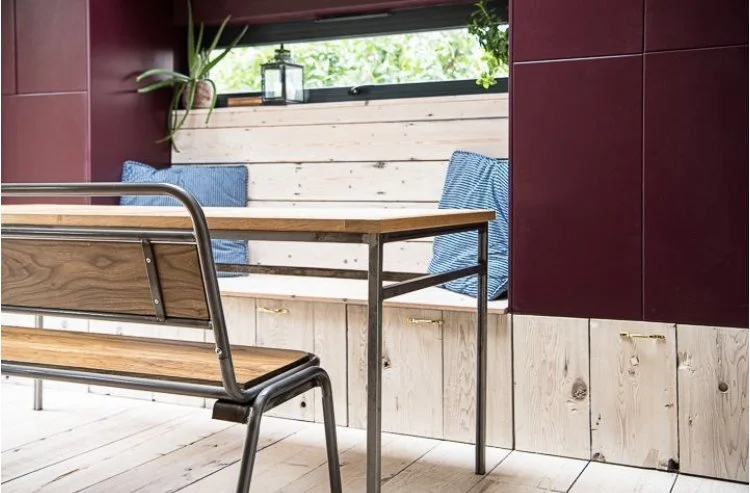 Interior of a modern room with a wooden table, metal-framed bench, wood-paneled wall, and window with greenery outside.
