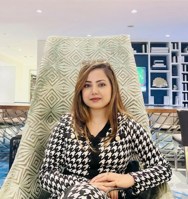 A woman with light brown hair and wearing a black and white houndstooth blazer sits in a high-backed, textured armchair in a modern, well-lit room with blue shelving and a TV in the background.