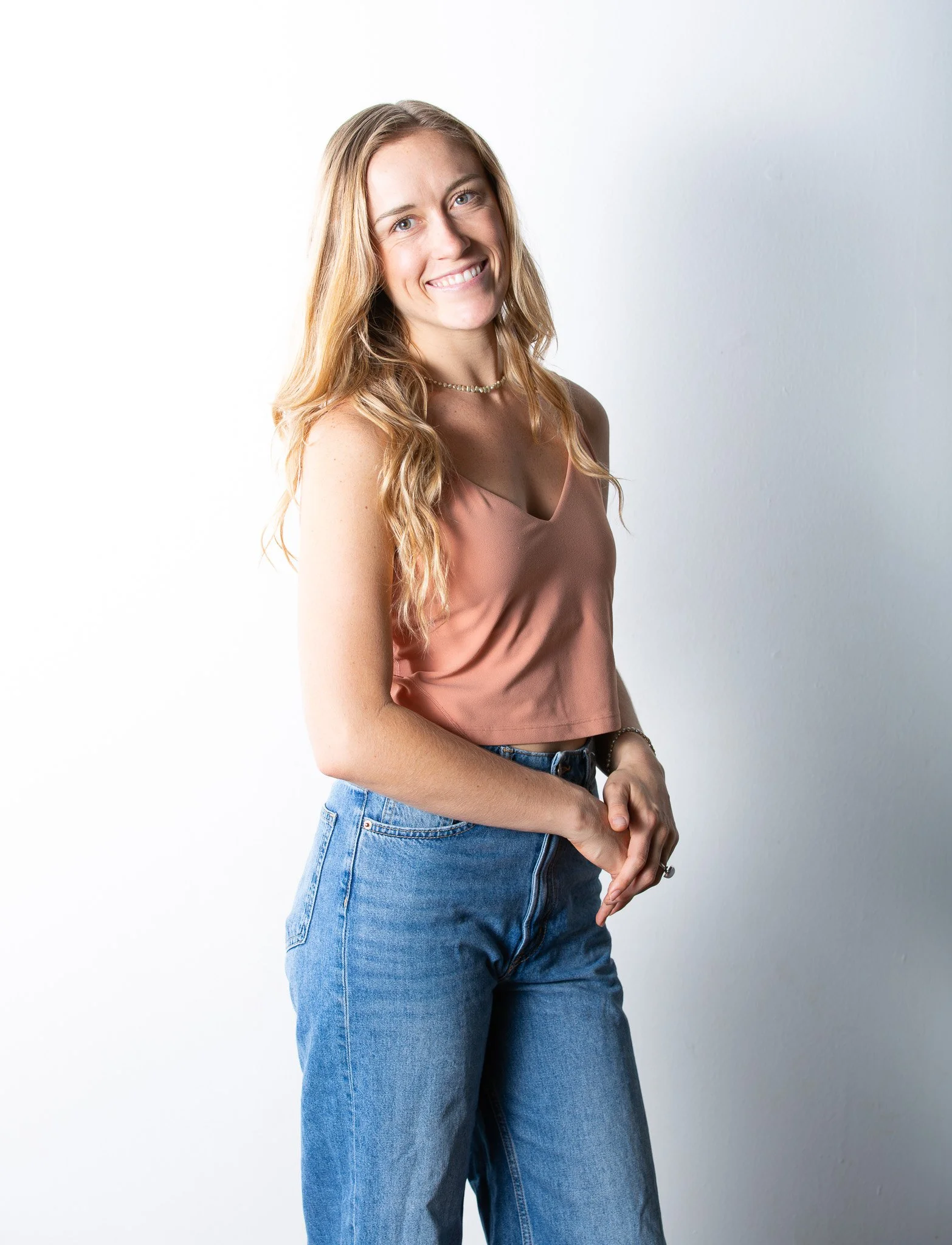 Young woman with long, wavy blonde hair, smiling, wearing a pink sleeveless top and high-waisted blue jeans, standing against a white background.
