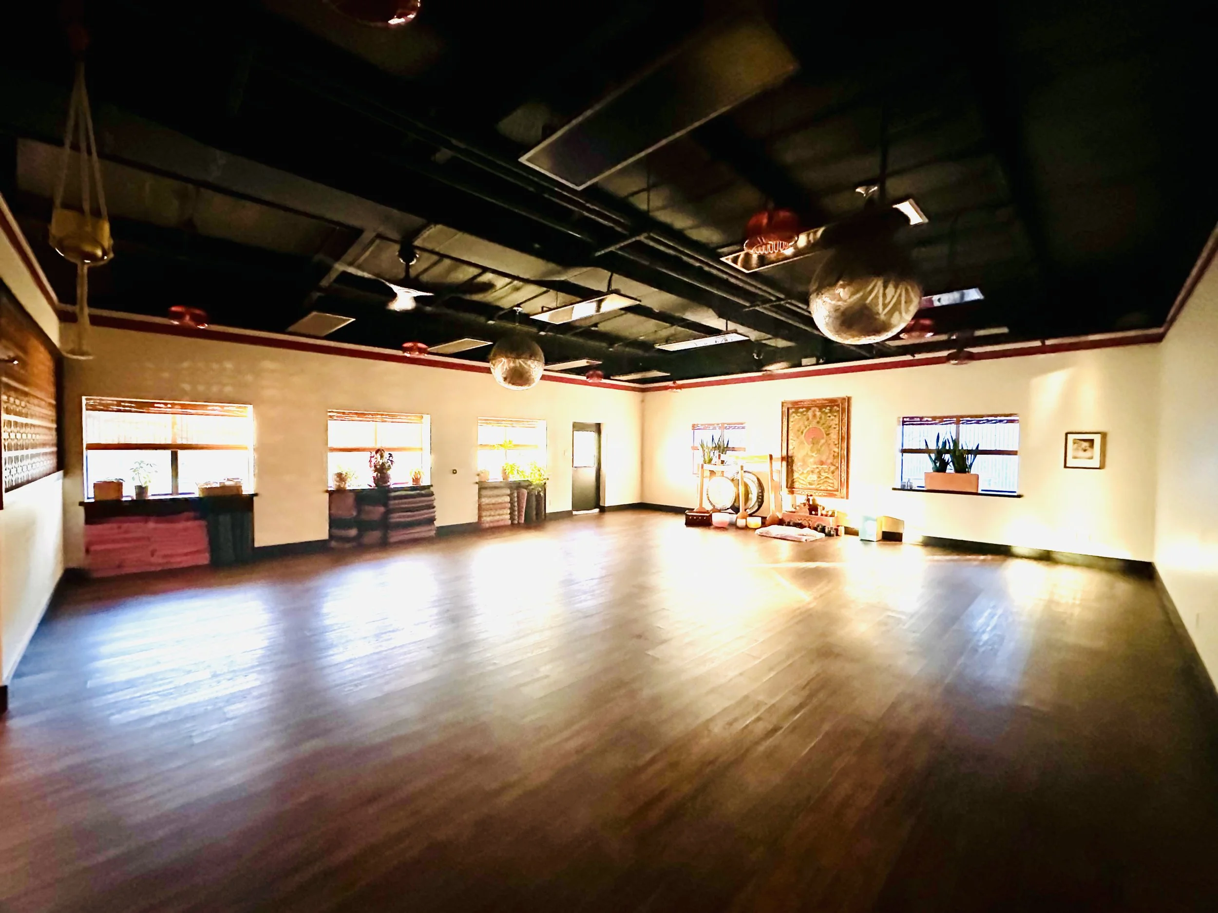 A spacious dance or yoga studio with wooden floors, large windows with blinds, and various decorative items and plants. The ceiling is black with visible pipes and hanging lamps.