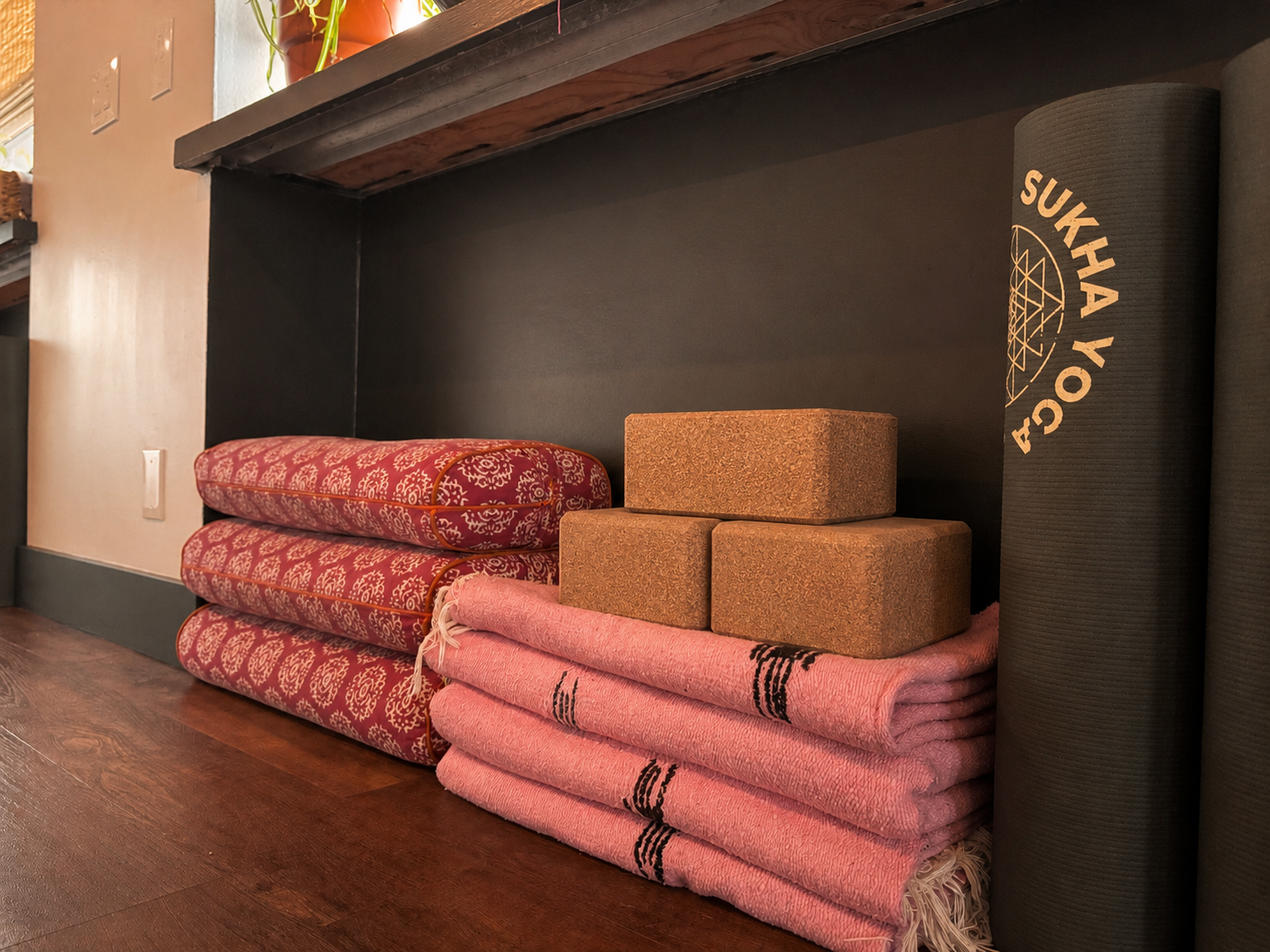Sukha Yoga branded yoga mats by Manduka rolled next to stacked colorful pink yoga bolsters, blankets and cork blocks stored in a forest green shelf  inside the Sukha Yoga Oltorf studio between South Lamar and 1st St in the 78704 neighborhood.