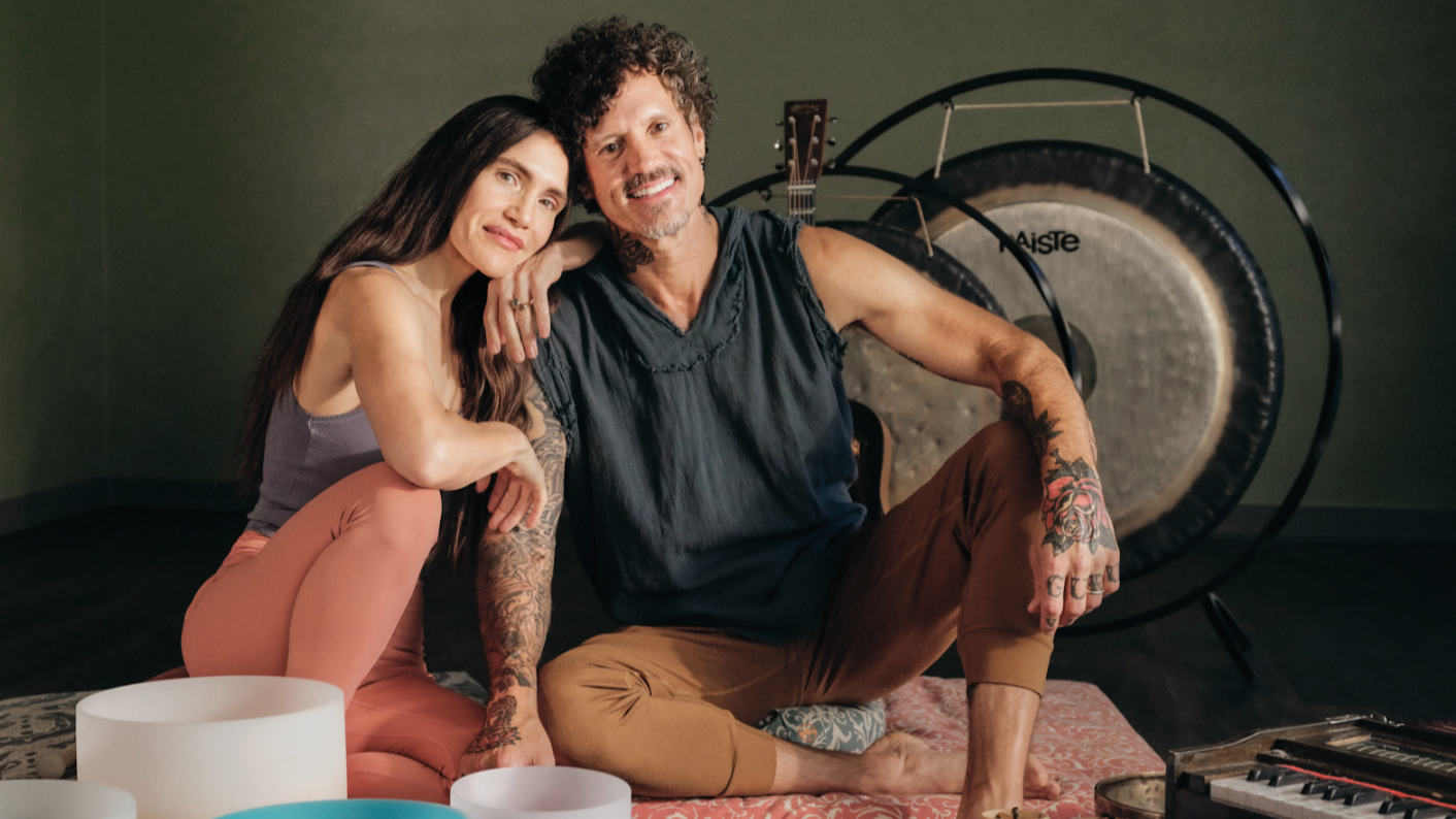 Erinn and Mark of Sukha Yoga Austin sit on the floor in their East Austin studio. There is a harmonium, crystal singing bowls and a large gong.