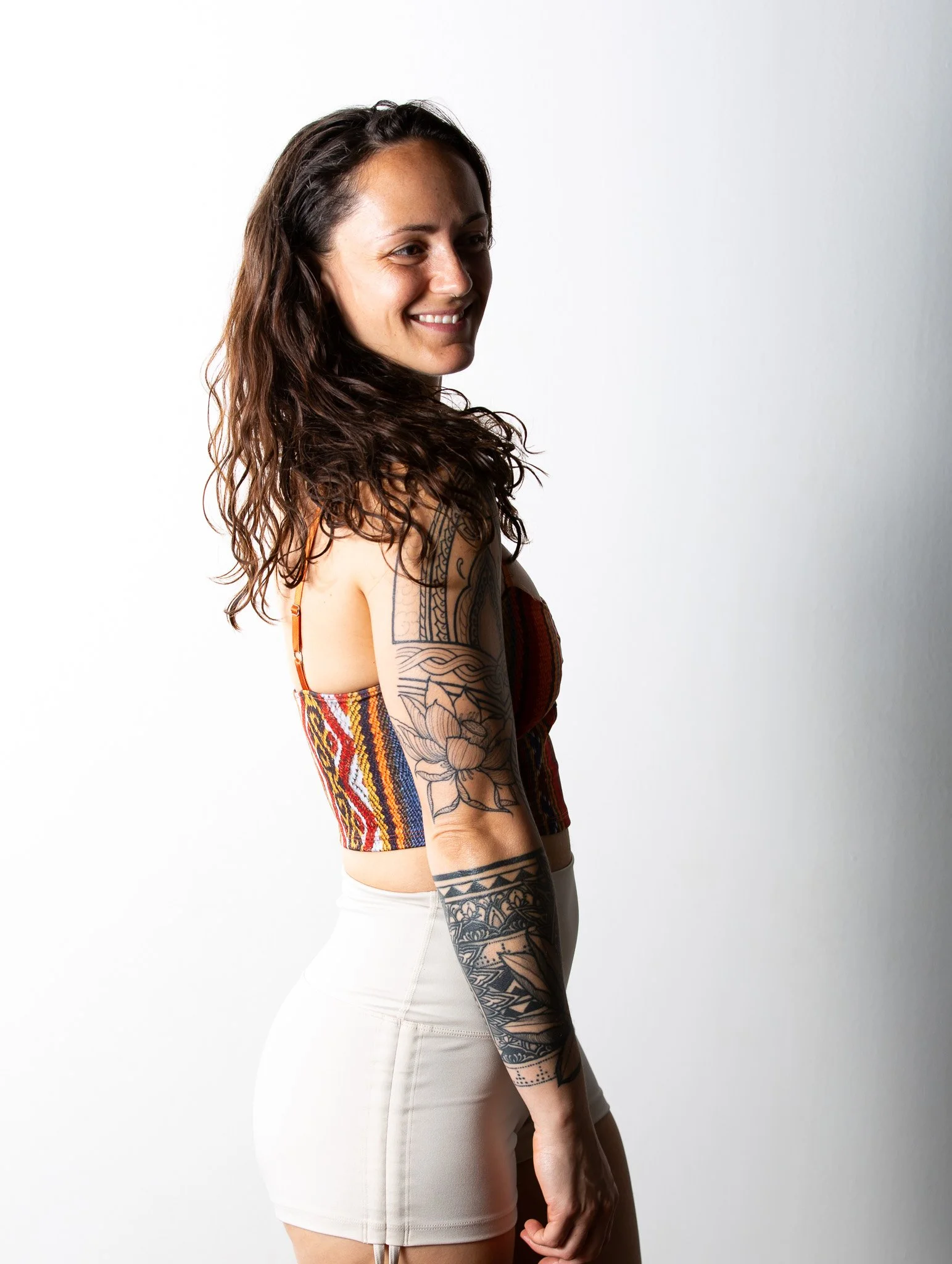 A smiling woman with long, wavy dark hair wearing a colorful, patterned crop top and white shorts, standing against a plain white background.