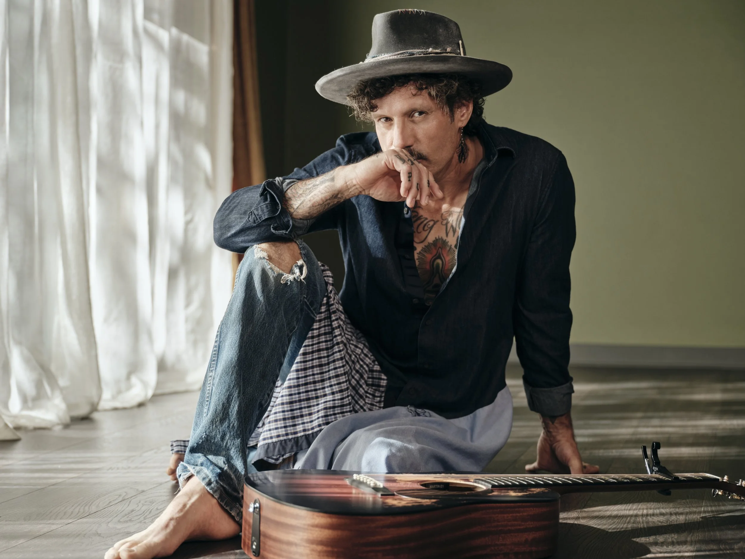 Mark Herron is sitting in Sukha Yoga East Austin studio wearing a custom made hat and dark clothing, is sitting on the floor with a guitar in front of him.