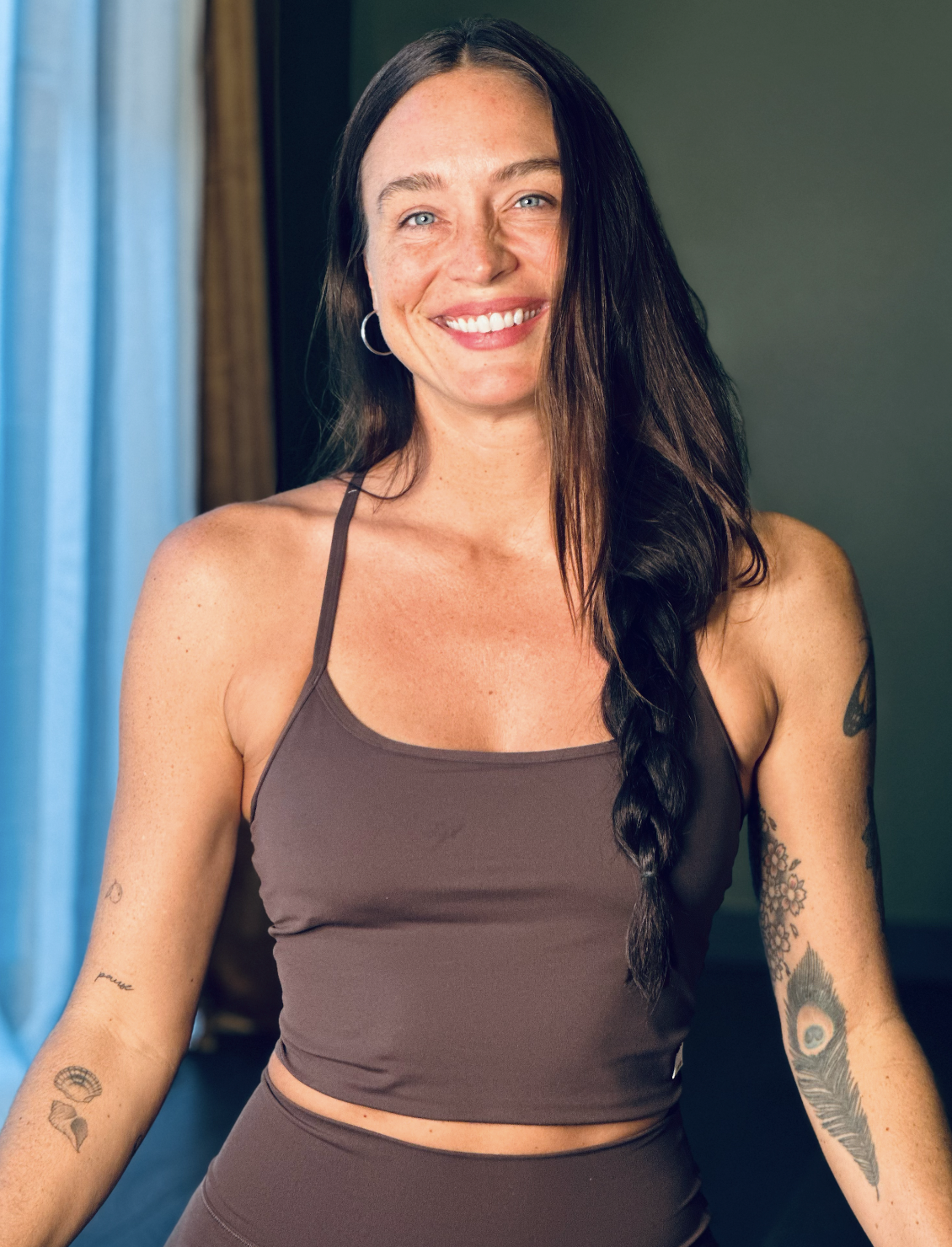 A smiling woman with long dark hair in a braid, wearing a brown tank top and earrings, standing indoors near a window with blue curtains.