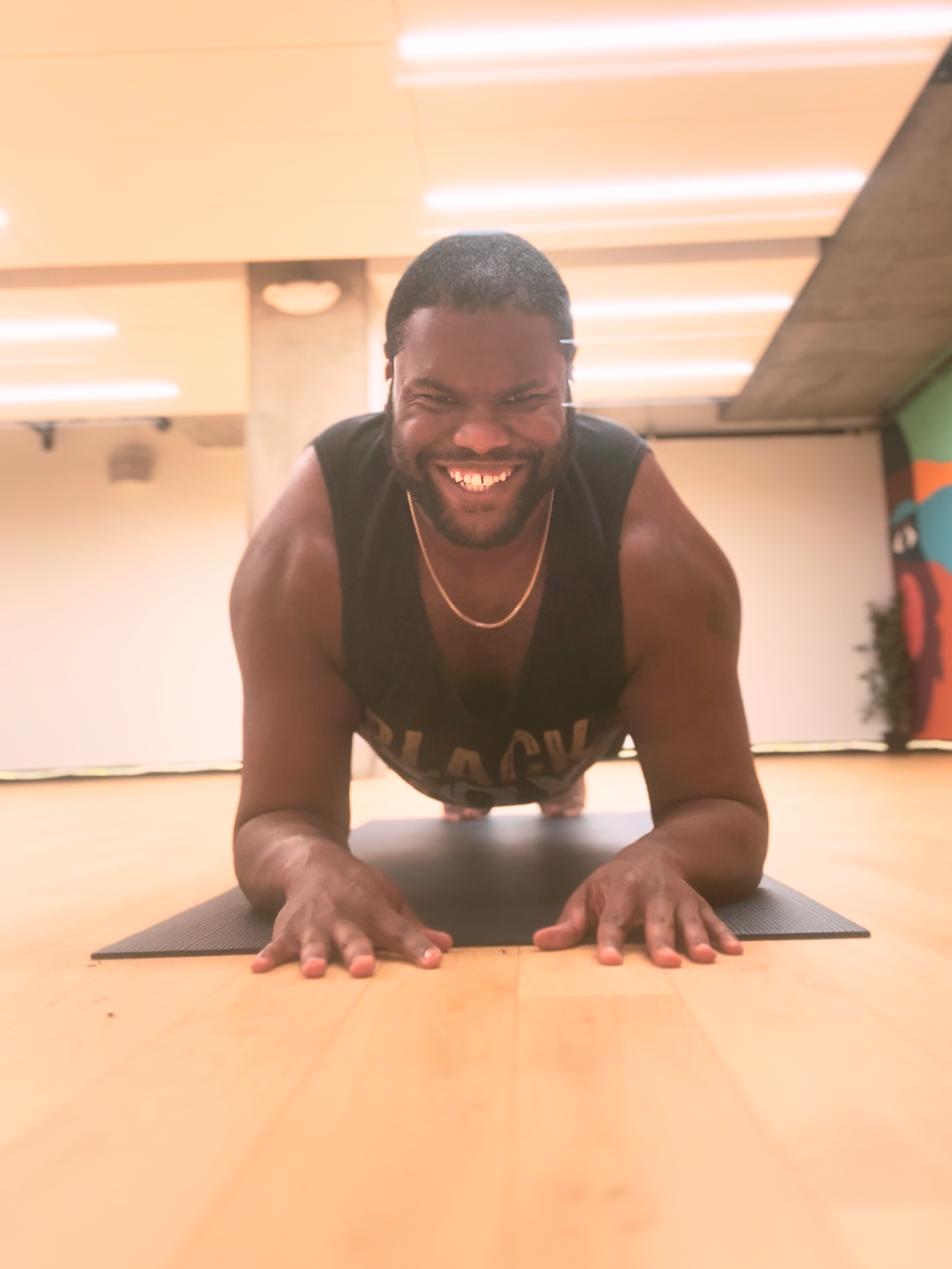 A man doing yoga in a plank position on a mat in a brightly lit indoor space, smiling at the camera.