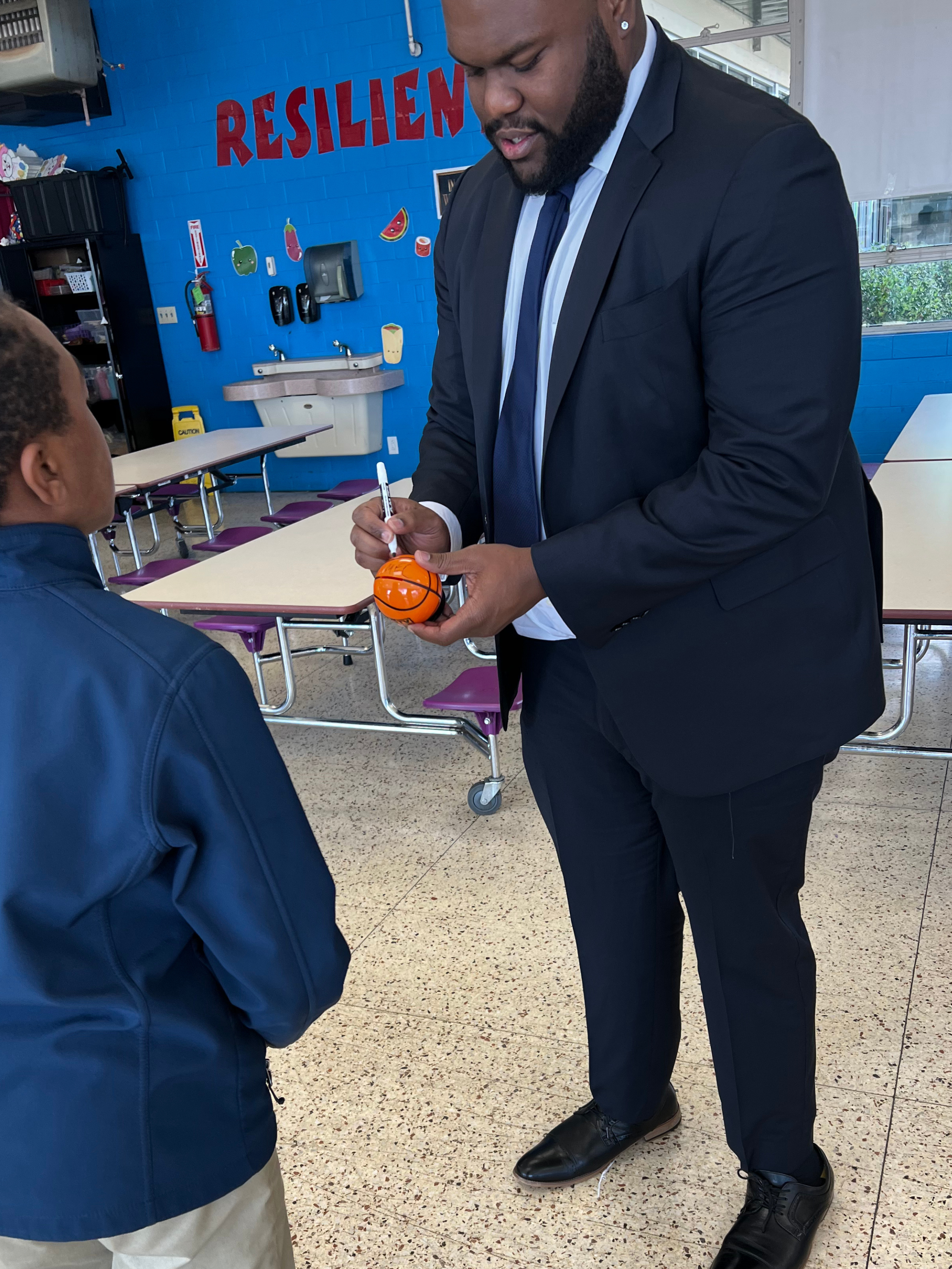 A man in a suit signing a basketball-shaped item held by a child in a school cafeteria.