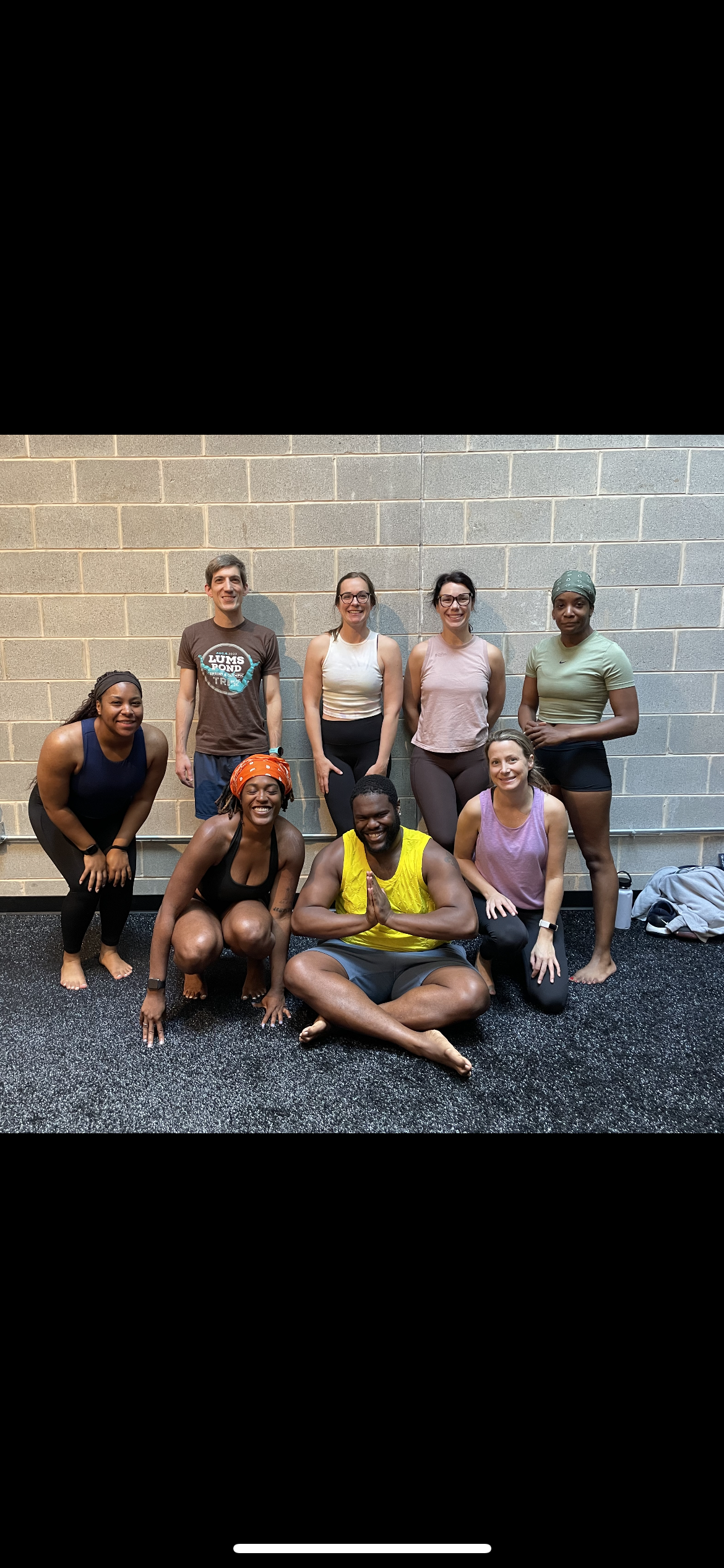 Group of nine diverse people in workout attire smiling and posing together in a fitness studio with a brick wall background.