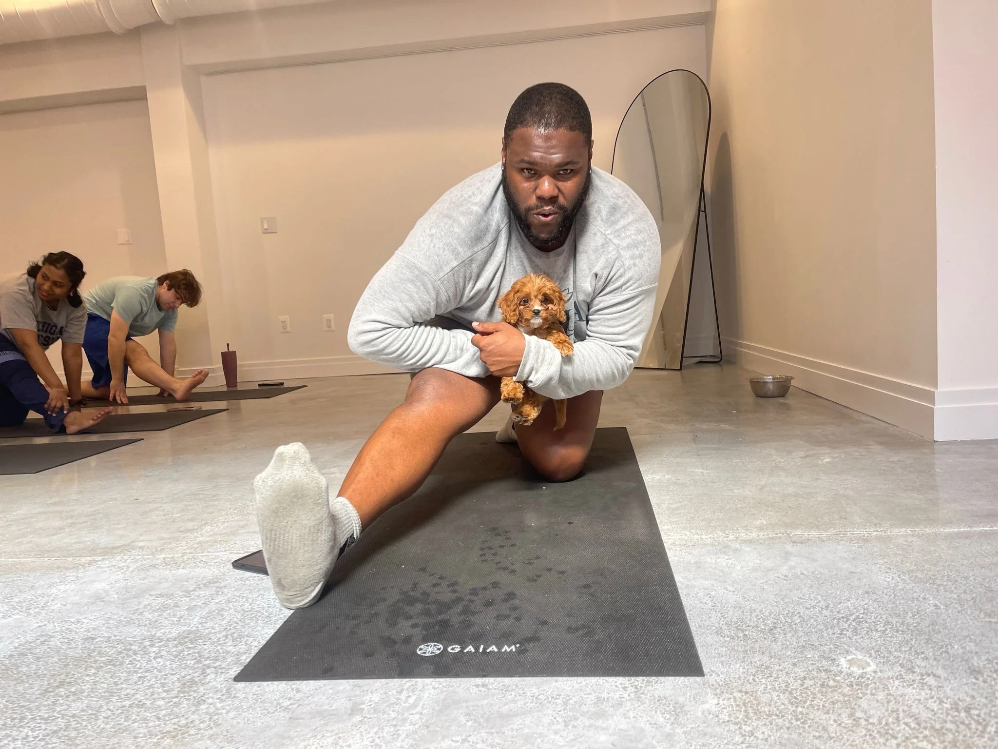 A man doing a yoga pose on a black mat while holding a small brown puppy, with two other people doing yoga in the background.