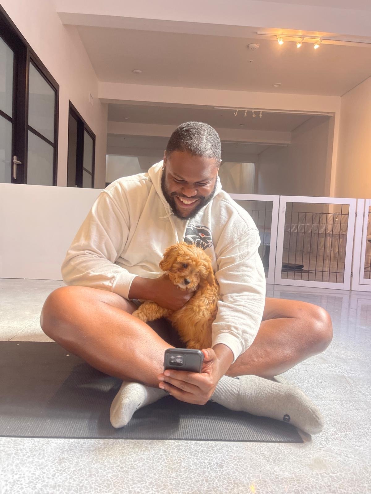 A man with a beard sitting cross-legged on the floor, holding a small, curly-haired brown puppy and looking at a smartphone, smiling.