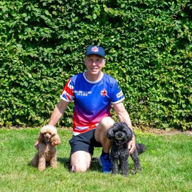 Jason and his dogs wearing a blue and red sports jersey and a matching cap kneels on grass with two small poodle dogs, one brown and one black, in front of a green leafy hedge.