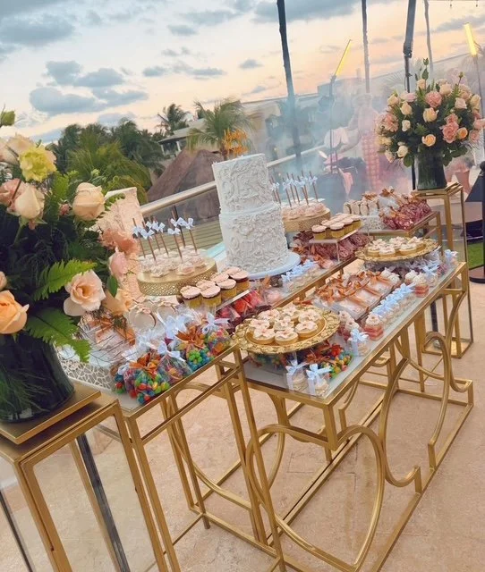 Dessert table with a white wedding cake, cupcakes, cookies, and colorful treats, decorated with pink and cream roses, located outdoors on a patio with a view of the sky and trees.