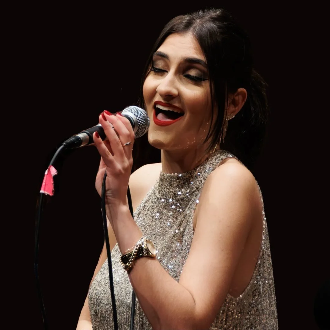 A woman singing into a microphone on stage, wearing a sparkling silver dress and red lipstick, with dark hair styled back, against a black background.