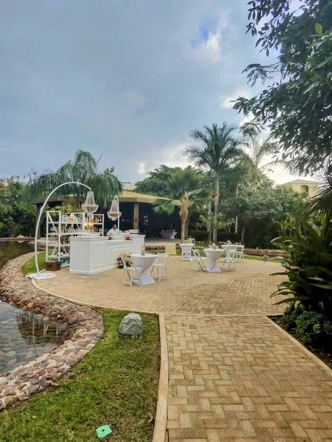Outdoor event setup with high-top table, chairs, and bar area surrounded by trees and landscaping, under a partly cloudy sky.