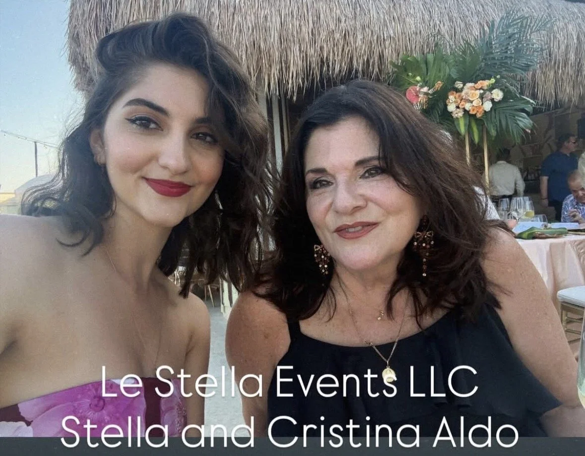 Two women sitting at an event, smiling at the camera, with floral decorations and a thatched roof background.