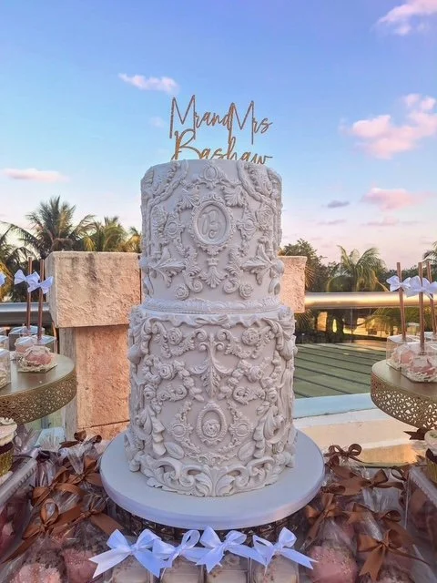 Two-layer white decorative cake with intricate floral and scroll patterns, topped with a gold sign reading 'Married Mrs. Bashaw' on a round white cake stand, outdoors with palm trees and a sunset sky in the background.