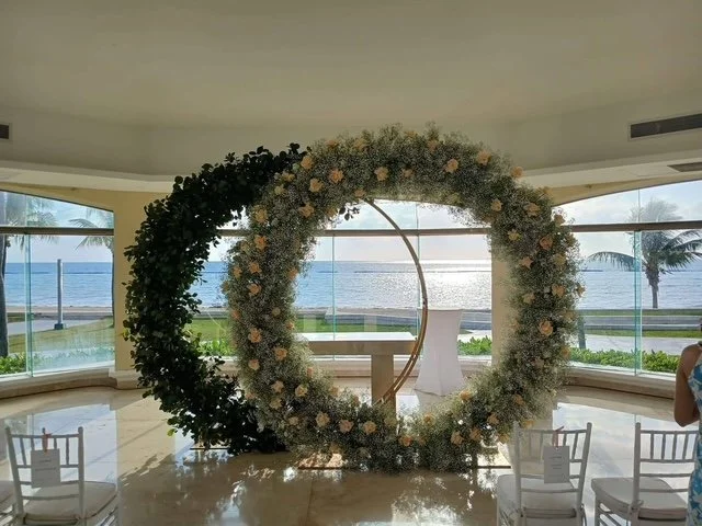 A large floral wreath with green and white flowers and leaves, arranged in a circle, positioned in front of a window with a view of the beach and ocean.