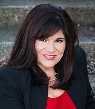 A woman with dark hair and light skin, smiling, wearing a black blazer and red top, sitting outdoors near stone steps.
