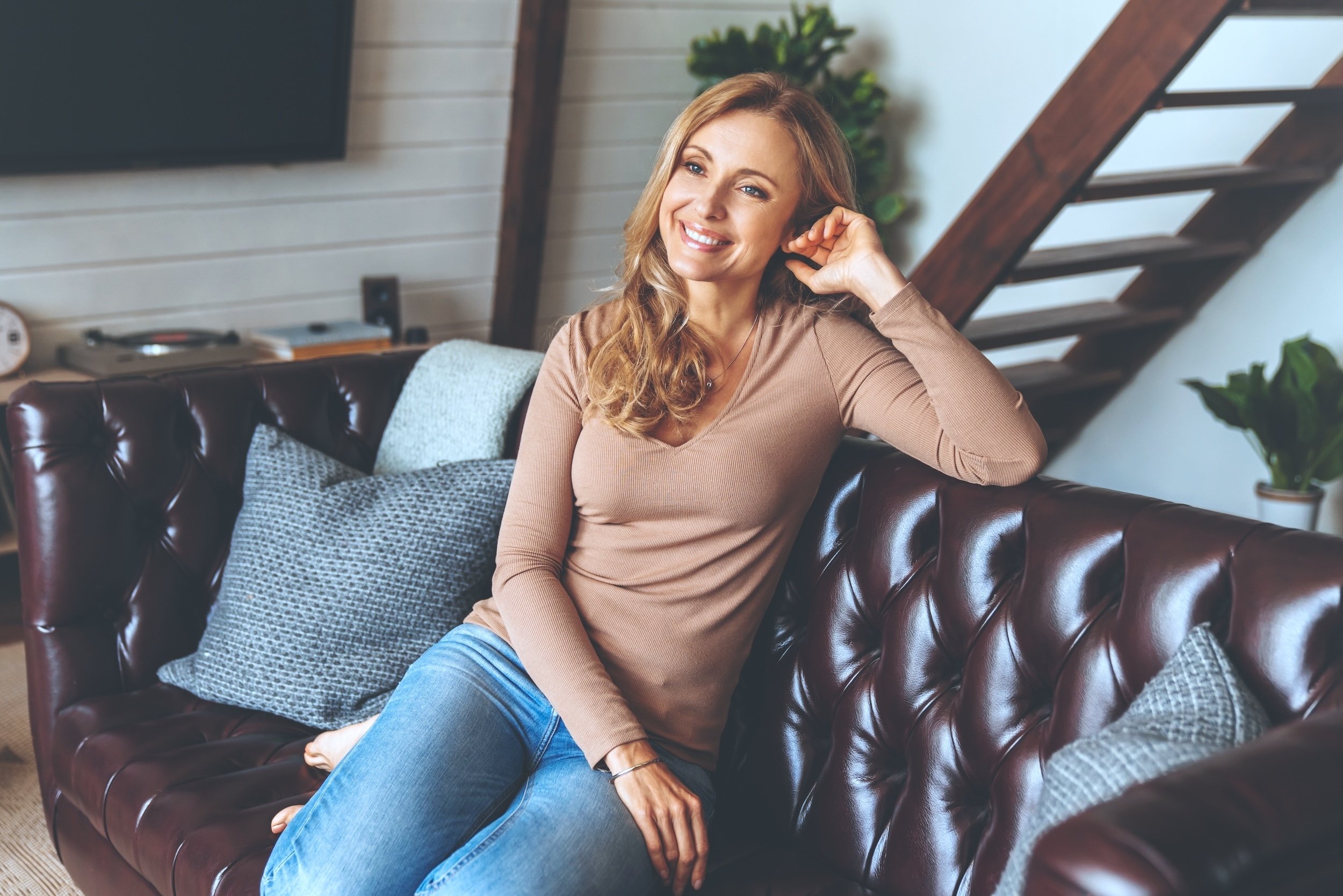 A woman with wavy blonde hair smiling while sitting on a brown leather couch in a cozy living room with a staircase and plants in the background.