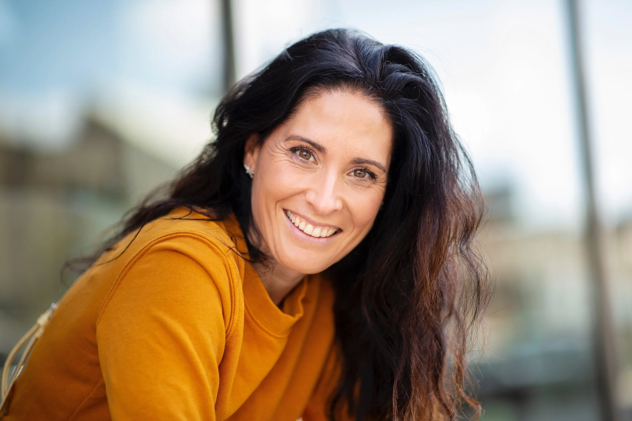 Smiling woman with long dark hair wearing a mustard yellow top, indoors with a blurred background.