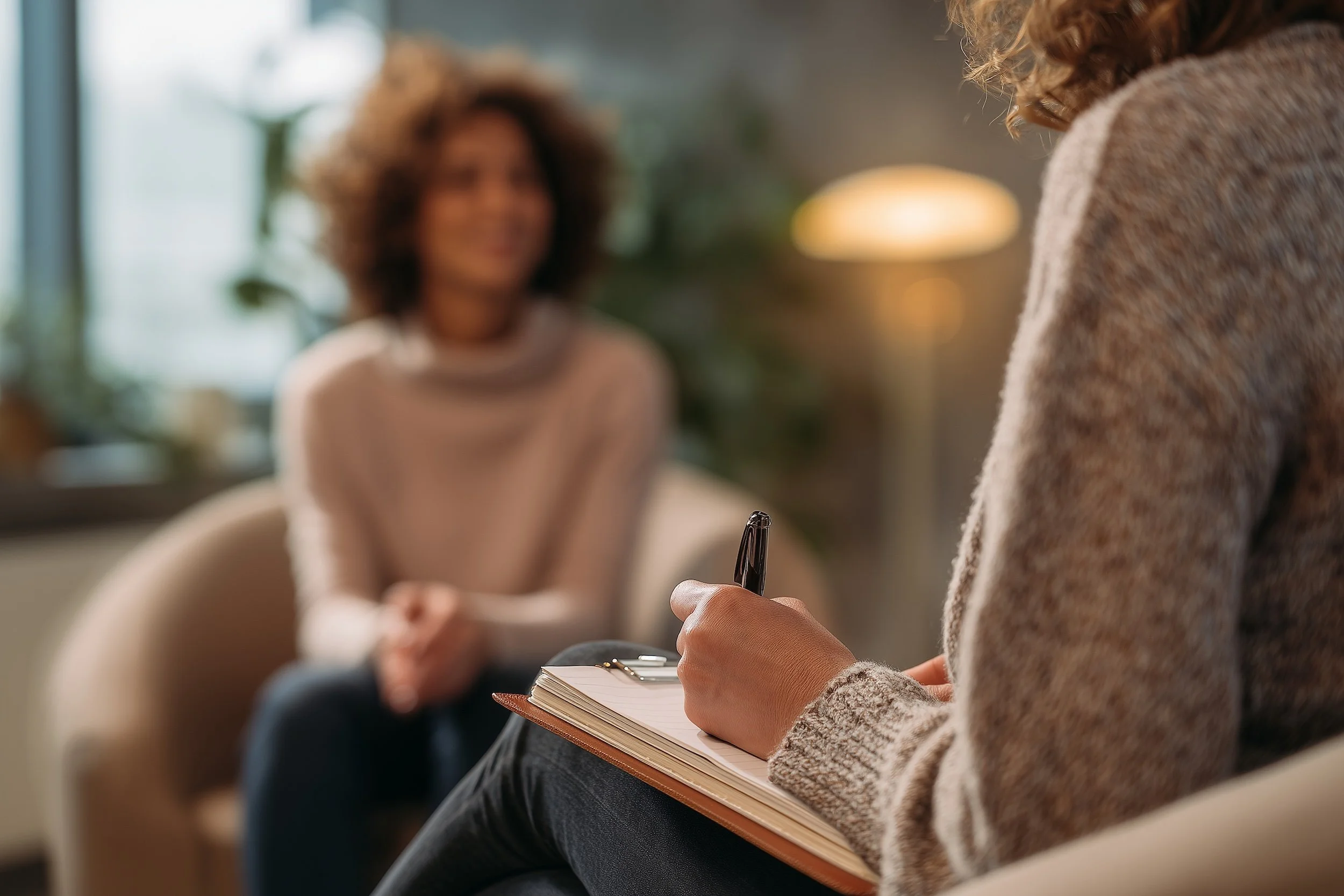 Two women having a conversation in a cozy, well-lit room; one woman is taking notes in a notebook, the other is smiling and sitting across from her.