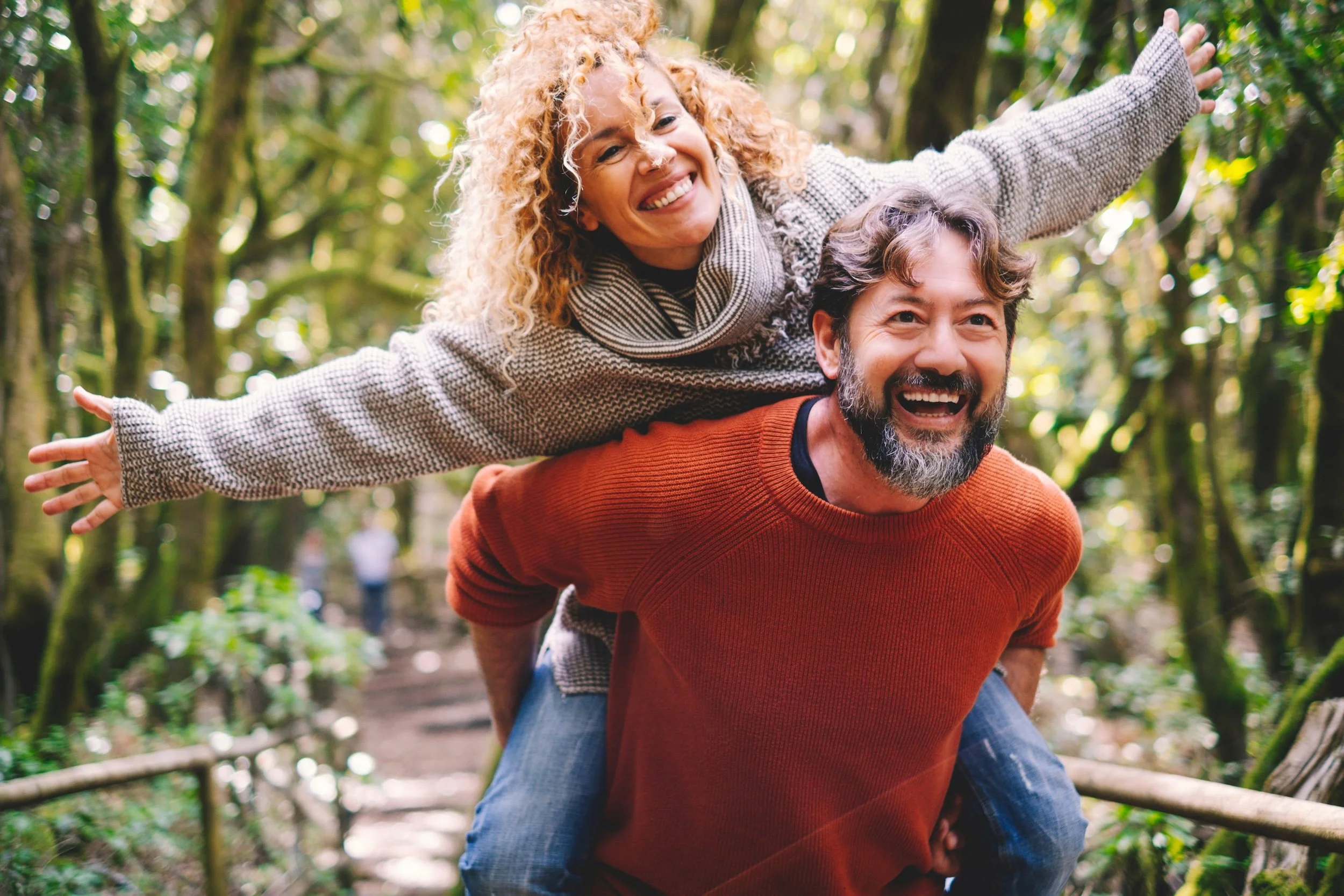 A joyful couple enjoying a walk in a forest, with the woman sitting on the man's shoulders and both smiling. The woman has curly blonde hair and is wearing a beige sweater, while the man has a beard and is wearing an orange sweater.