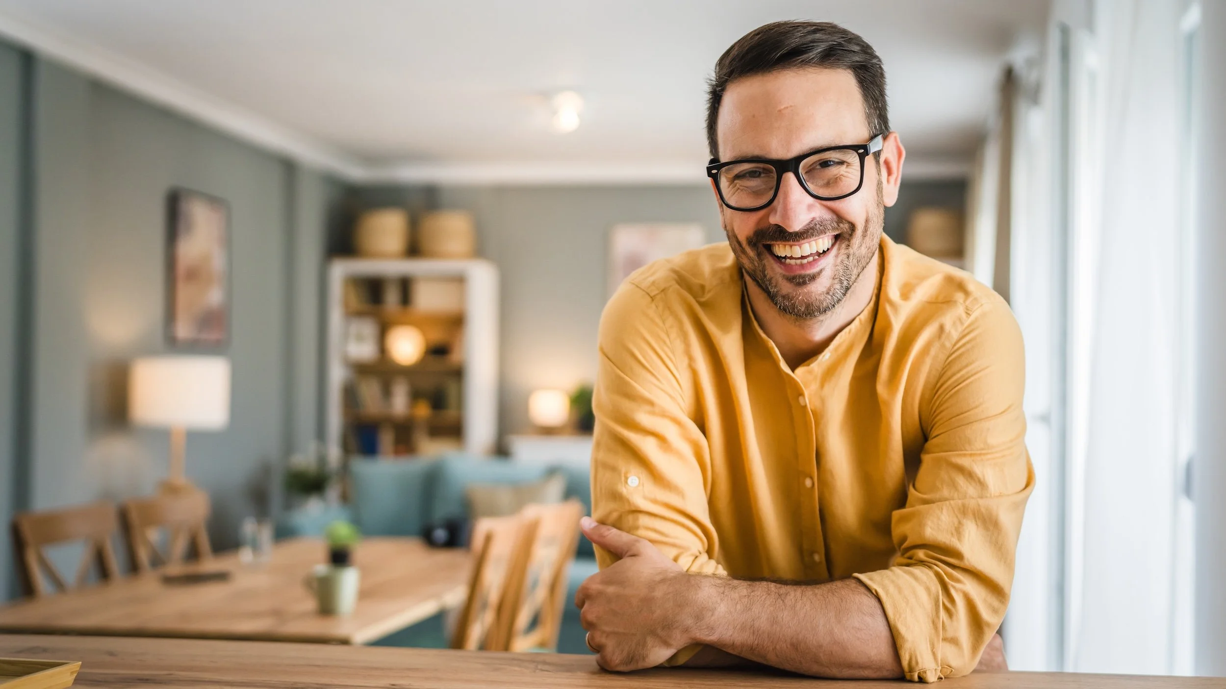 A smiling man with glasses and a beard leaning on a wooden counter inside a well-lit living room, with a dining table and bookshelf in the background.