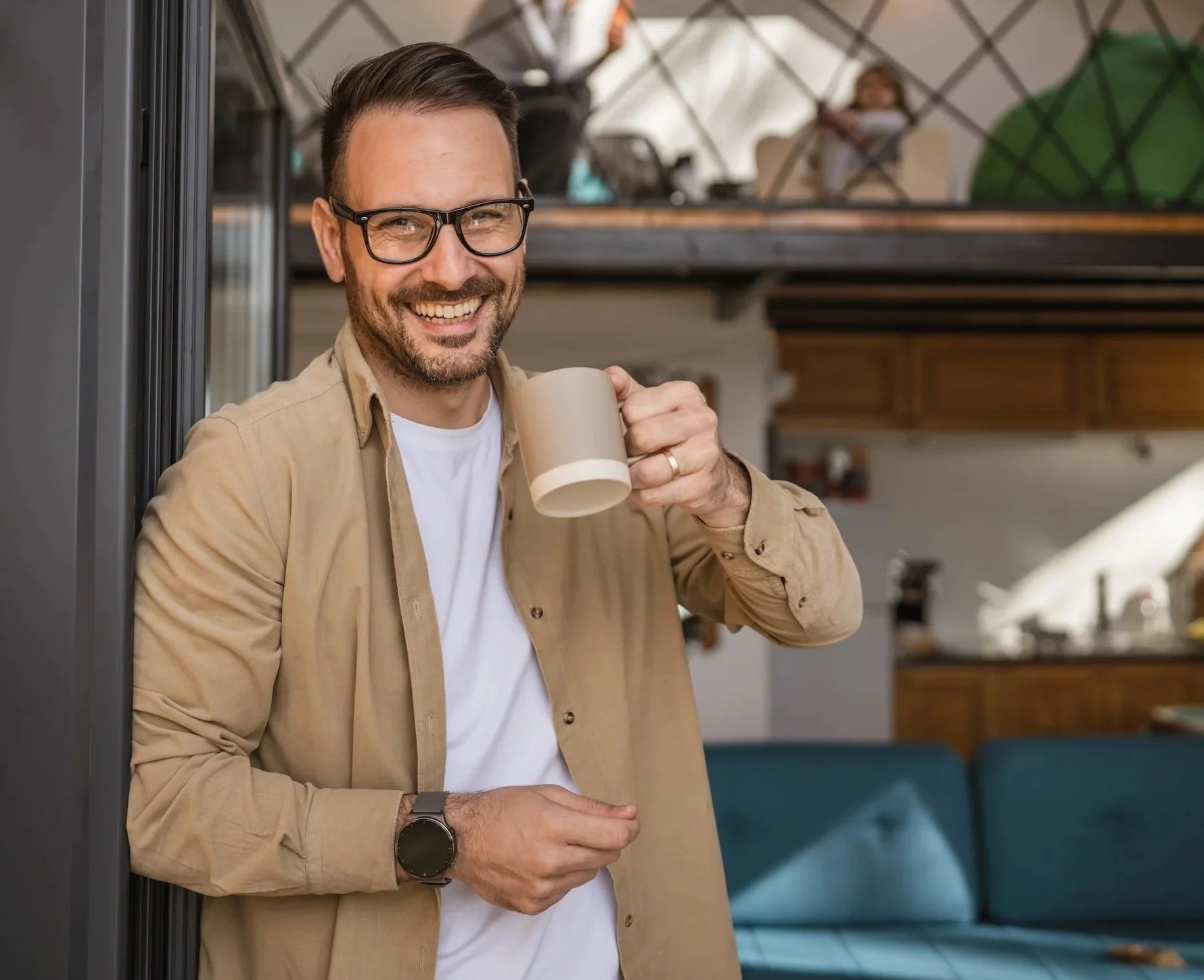 A man with glasses smiling and holding a beige coffee mug in a cozy, modern kitchen or cafe with wooden cabinets and a teal sofa in the background.